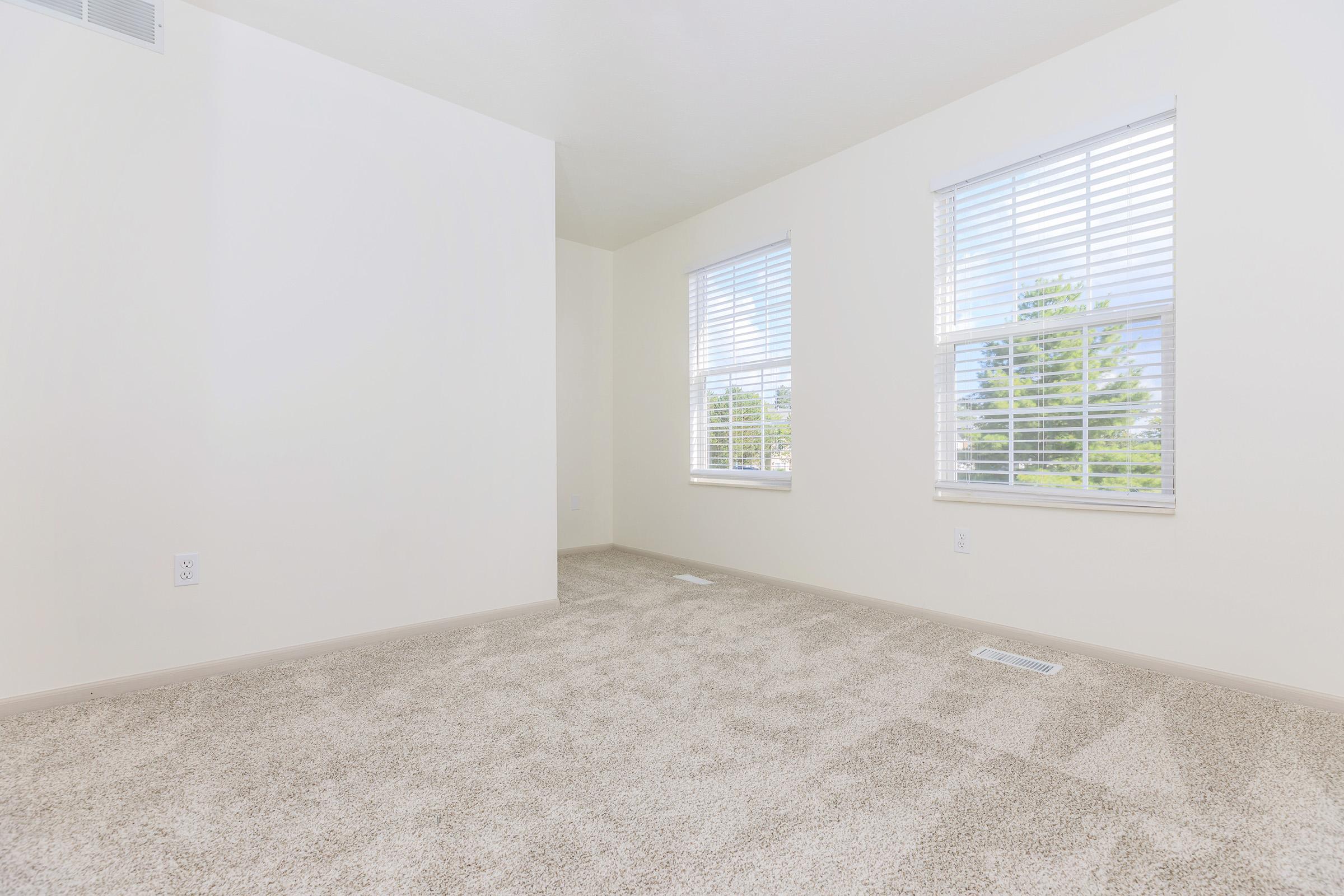 A vacant room with light-colored walls and carpet. Two windows with horizontal blinds allow natural light to enter, illuminating the space. The room features a plain design, with no furniture, creating an open and airy atmosphere.