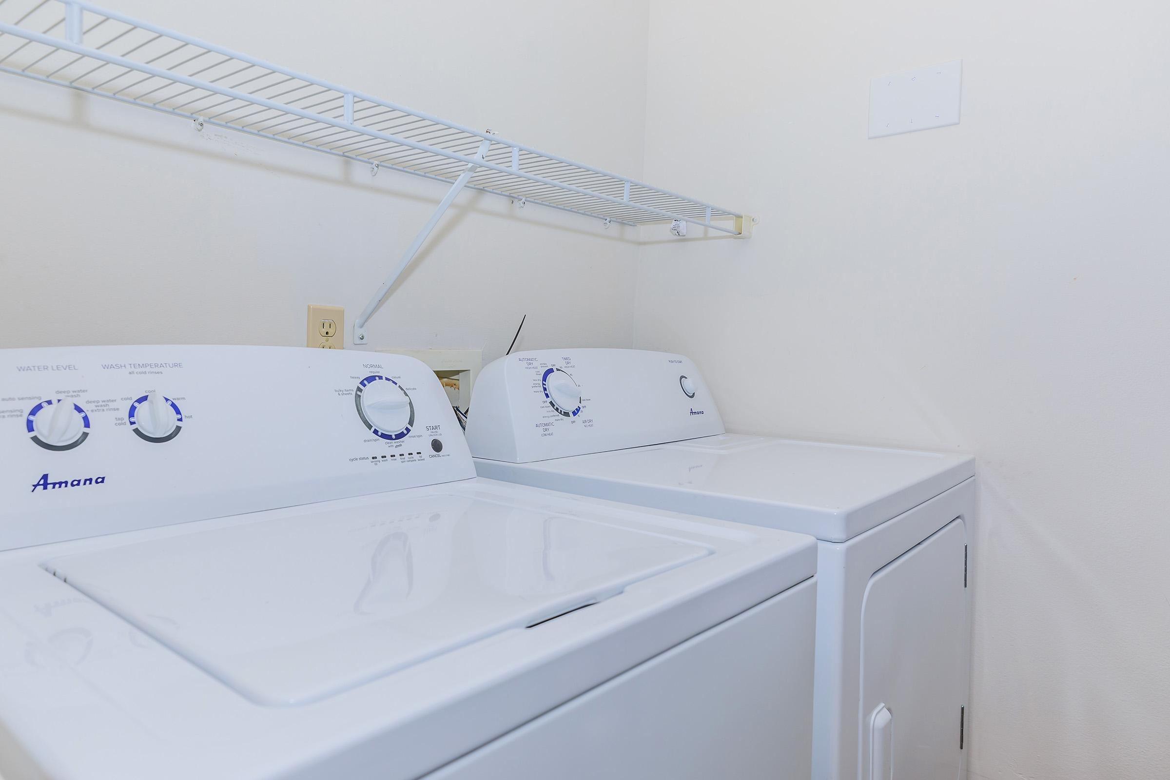 A clean laundry room featuring a white washer and dryer, positioned side by side against a light-colored wall. Above the appliances, there is a simple wire shelf for storage. The space is well-lit and organized, ideal for laundry tasks.