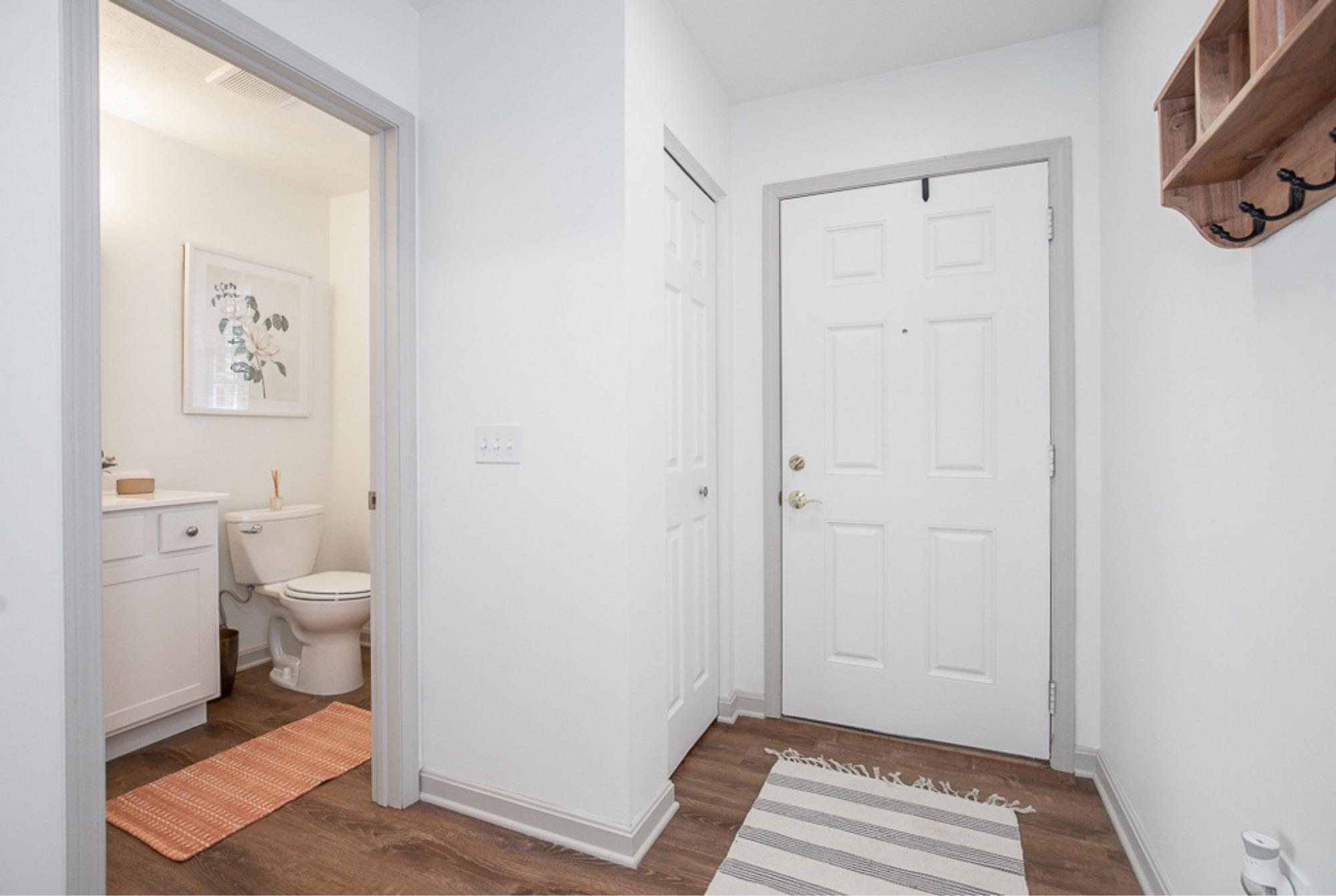 A bright entryway featuring a white door, a small bathroom visible on the left, and a coat rack on the wall. The floor is decorated with a striped area rug and a textured mat. Light-colored walls create an open, inviting atmosphere.