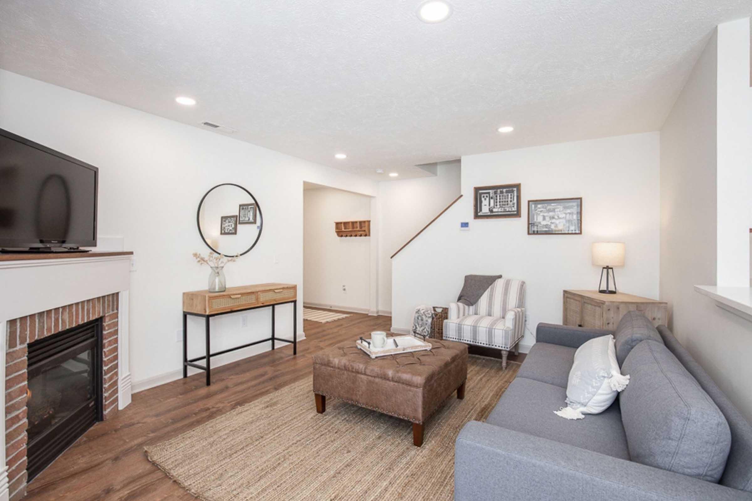 A modern living room featuring a cozy gray sofa, a brown upholstered ottoman, and a light-colored armchair. A flat-screen TV is mounted on the wall above a brick fireplace. Decorative items are displayed on a side table, and a round mirror hangs nearby. The space is bright and inviting with wooden flooring.