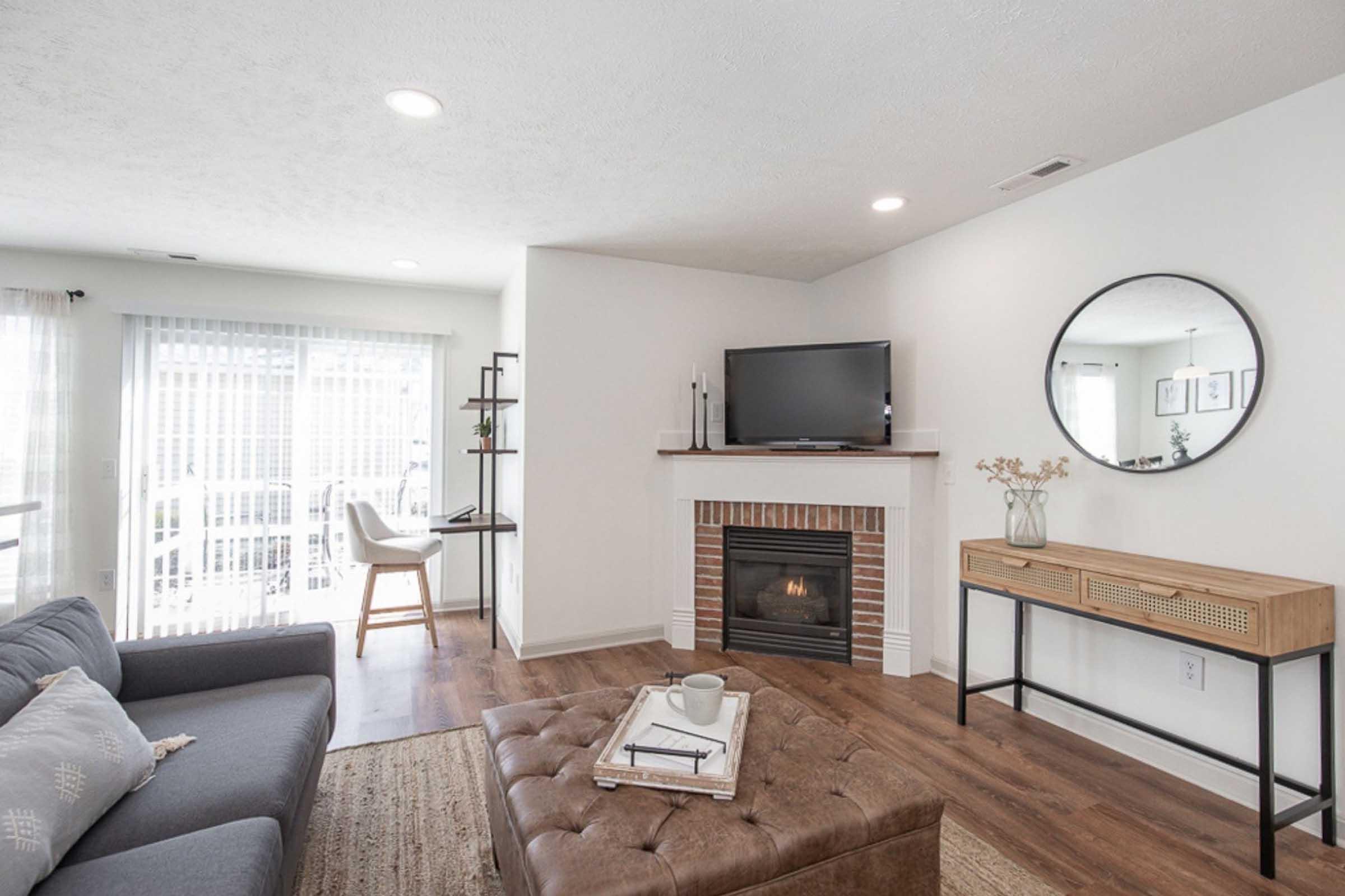 A modern living room featuring a gray sofa, a round mirror above a wooden console table, and a flat-screen TV mounted on the wall. A cozy fireplace is integrated into the decor, and a light-colored rug is placed on a wooden floor. Natural light filters through sheer curtains, illuminating the space.