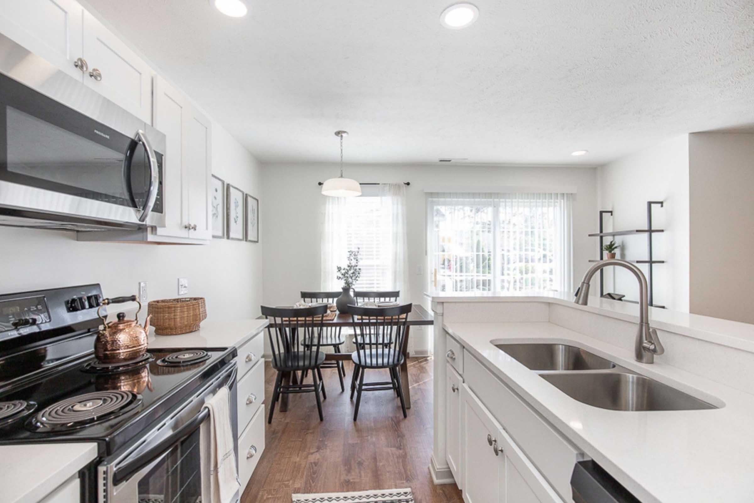 Modern kitchen with white cabinets, a black stove and microwave, and stainless steel sink. Nearby, there’s a dining area with a wooden table and black chairs. Large windows allow natural light, and decor includes wall art and a simple centerpiece on the table. The floor is hardwood.