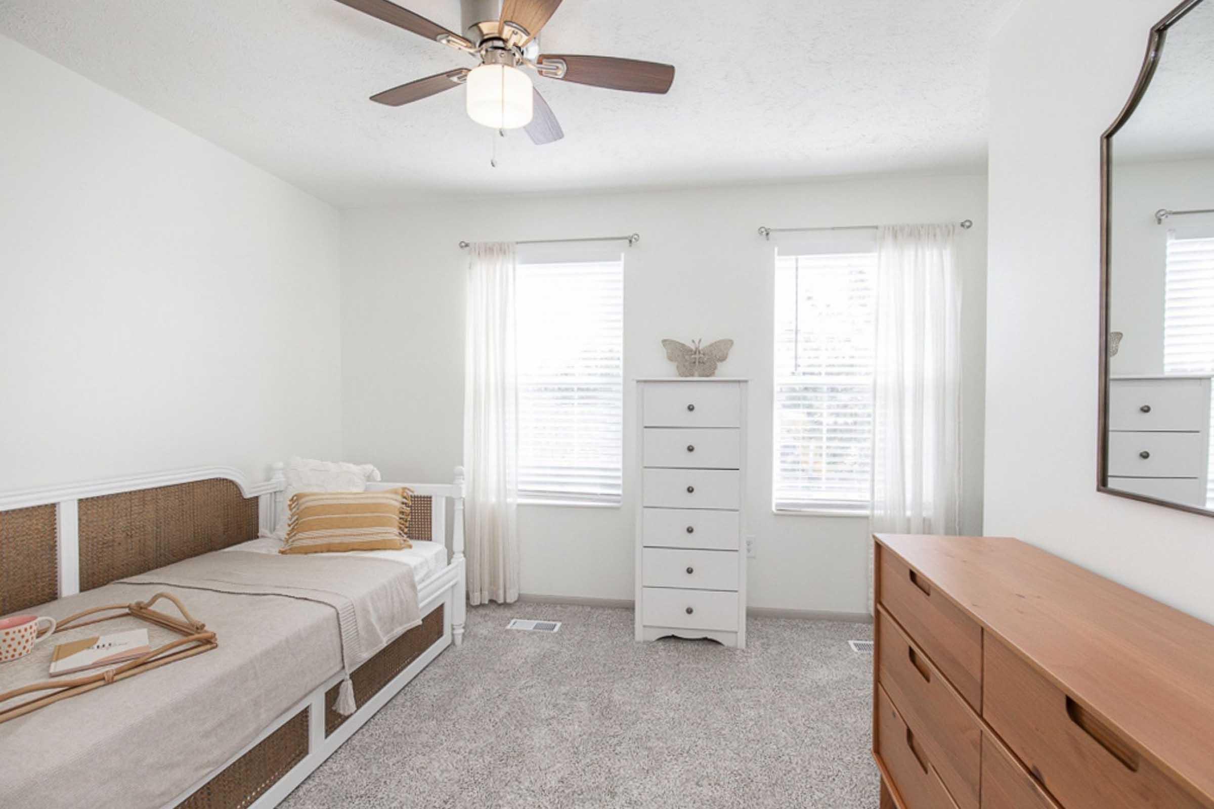 A light and airy bedroom featuring a single bed with neutral bedding, a decorative throw pillow, and a bedside table. There’s a white dresser with six drawers, a large mirror on the wall, and two windows dressed with sheer curtains, allowing natural light to brighten the space. The floor is carpeted.