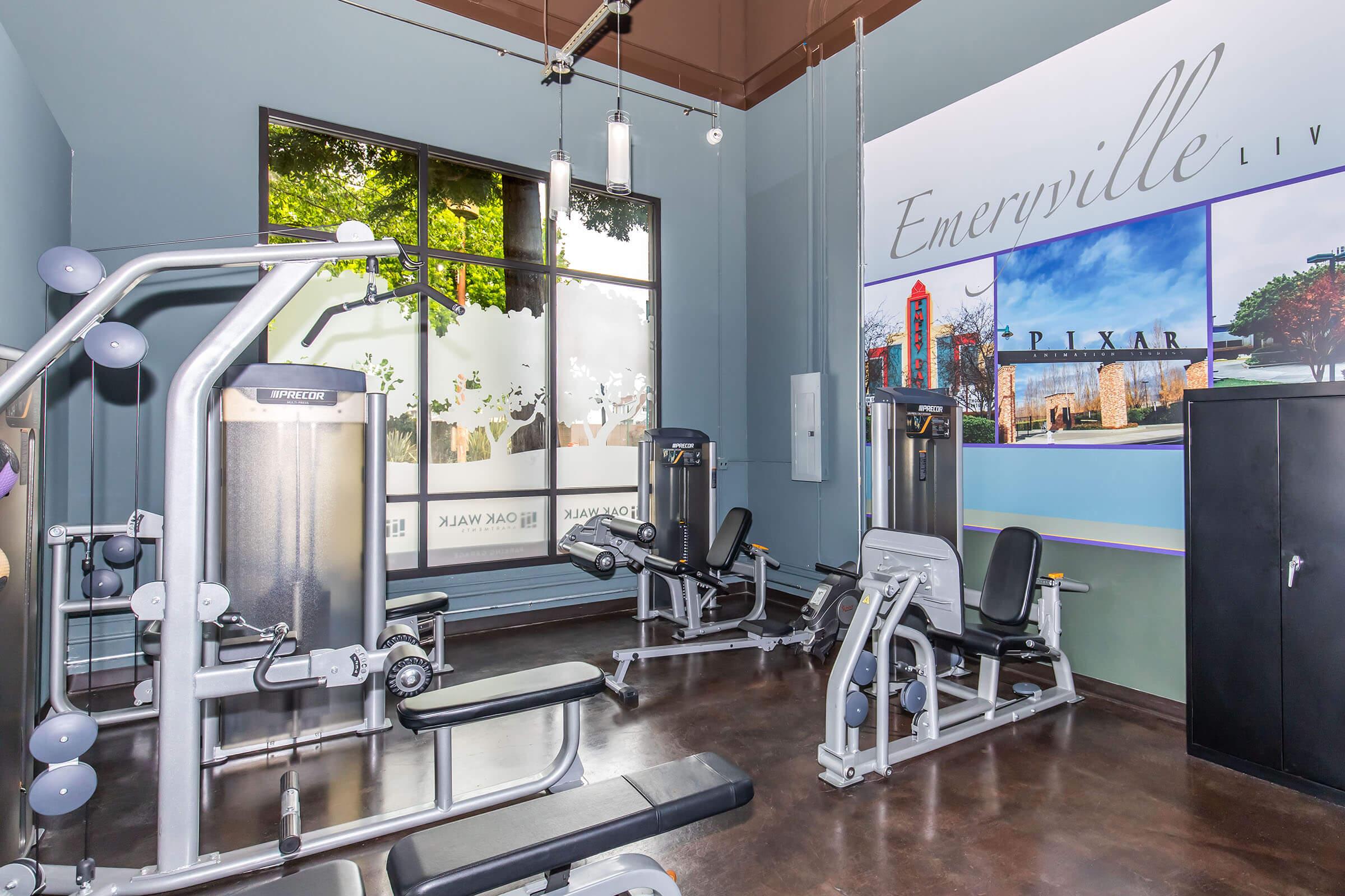 A modern gym interior featuring exercise equipment like weight machines and benches, with large windows that let in natural light. A wall mural showcasing landmarks of Emeryville and Pixar can be seen in the background, enhancing the ambiance of the fitness space.