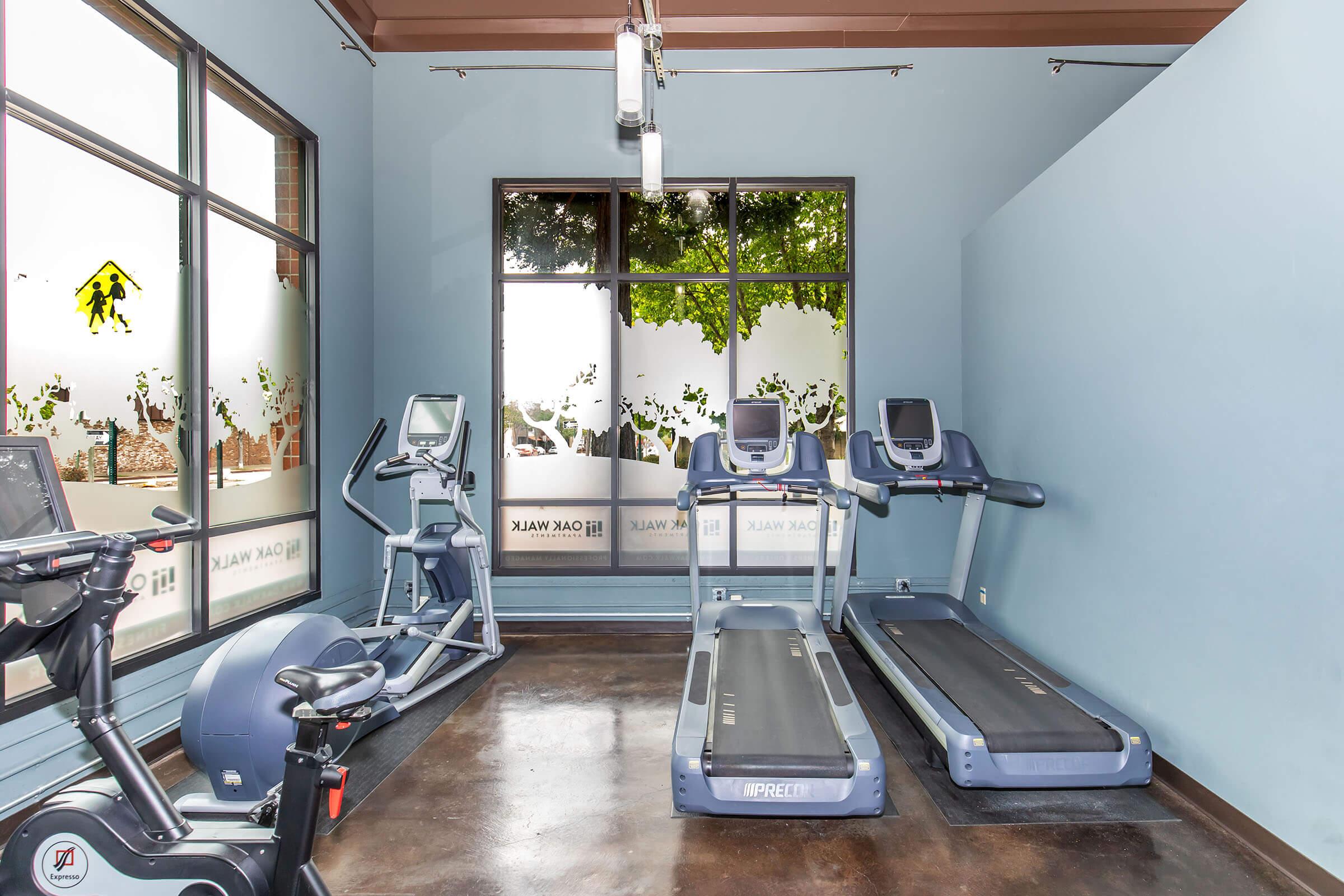 A fitness room featuring two treadmills and a stationary bike. The room is well-lit with large windows showcasing outdoor greenery. The walls are painted light blue, creating a calm atmosphere for exercise.