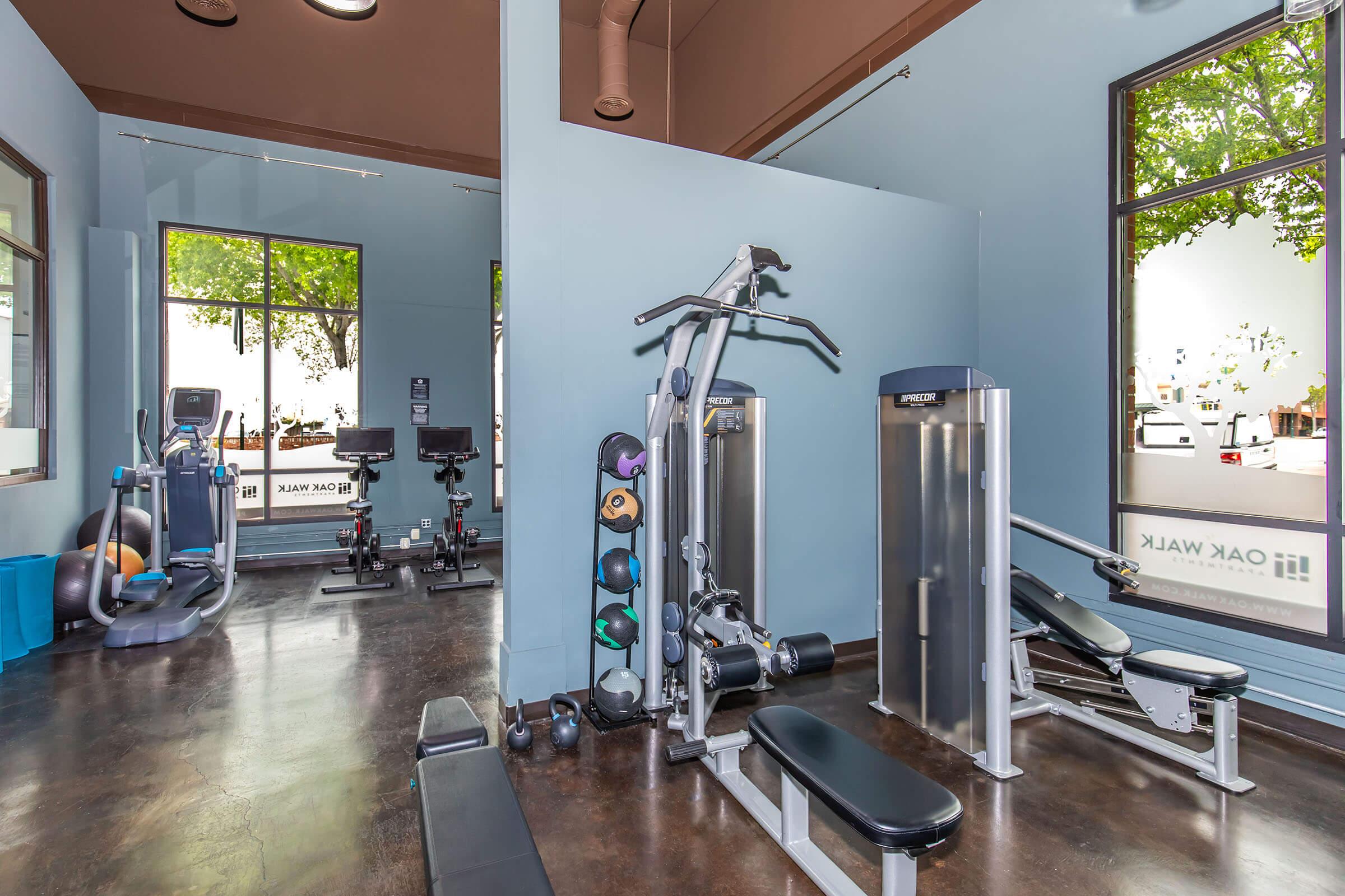 A bright and clean gym interior featuring various fitness equipment. On the left, there’s a stationary bike and weights, while a multi-station exercise machine and an elliptical trainer are in the center. Large windows allow natural light to fill the space, enhancing the inviting atmosphere.