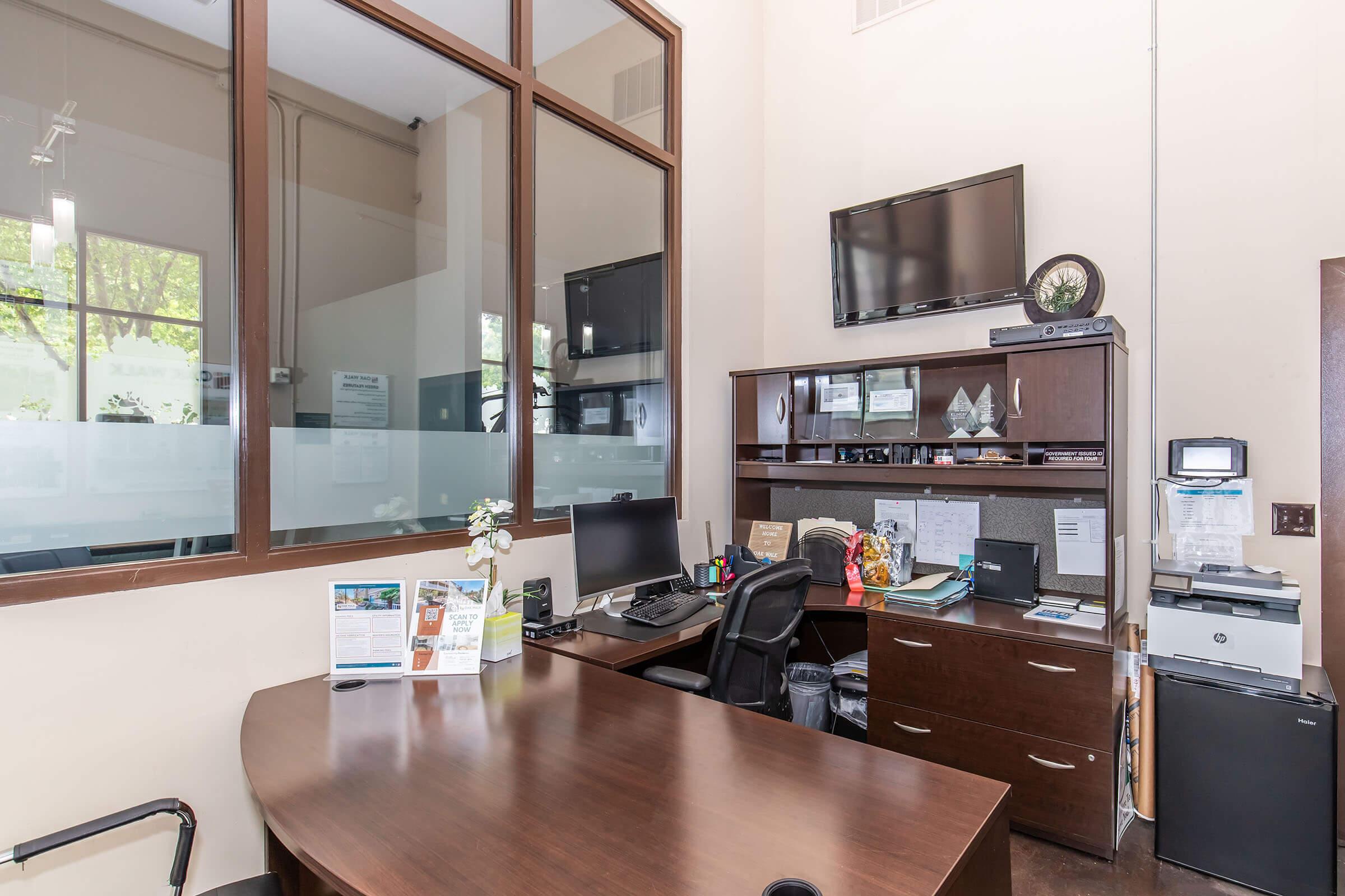 A modern office space featuring a large wooden desk with a black office chair, a computer monitor, and a printer. There is a wall-mounted television, large windows allowing natural light, and a bookshelf with various documents and decorative items. The overall ambiance is professional and organized.