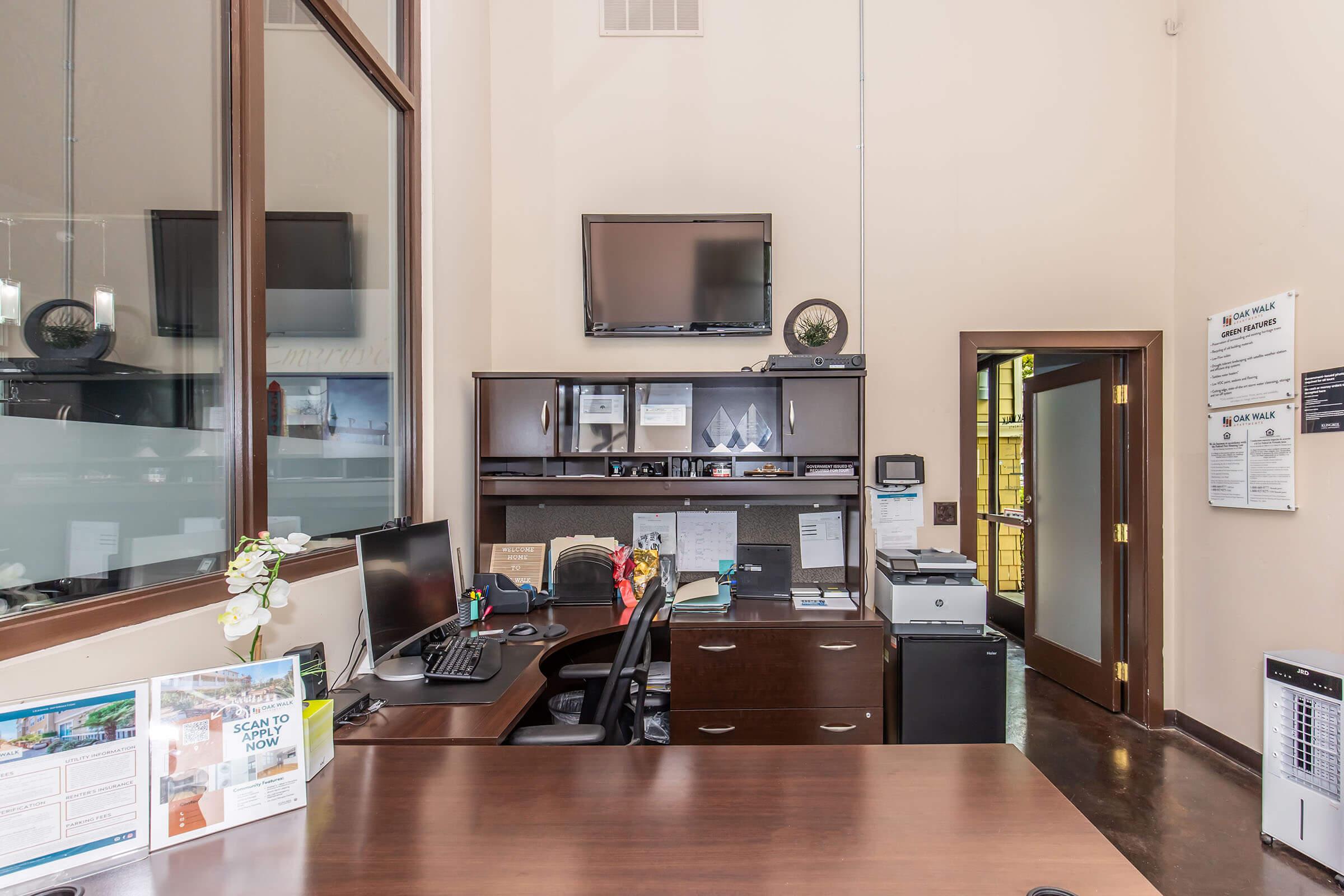 A modern office reception area featuring a dark wooden desk, a computer setup with monitors, and a printer. Wall-mounted TV is visible, along with organized paperwork and decorative items. Large windows provide natural light, creating a professional and welcoming atmosphere.