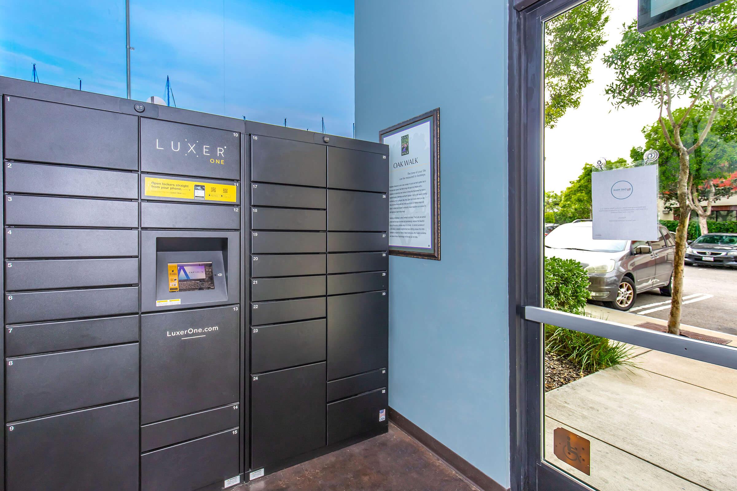 A black parcel locker system titled "LUXER ONE" is positioned near a glass door. The locker features a digital screen and various compartments for package storage. Outside, parked cars and greenery can be seen through the door. A sign with instructions is also present on the wall nearby.