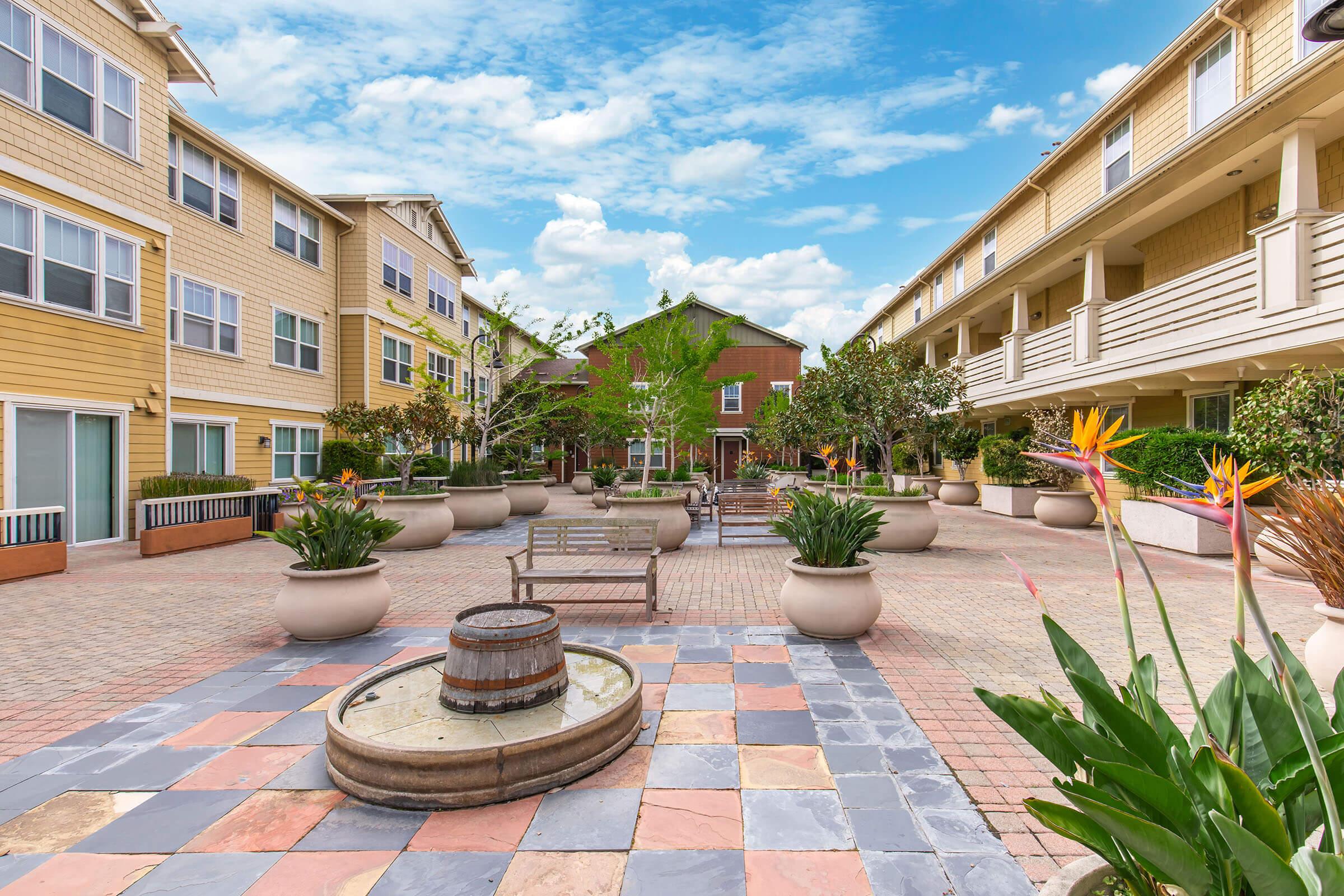 A landscaped courtyard with potted plants and flowers, featuring a central fountain and stone seating areas. Surrounding buildings are two to three stories high with a mix of yellow and neutral colors, and green trees provide shade against a blue sky with clouds.