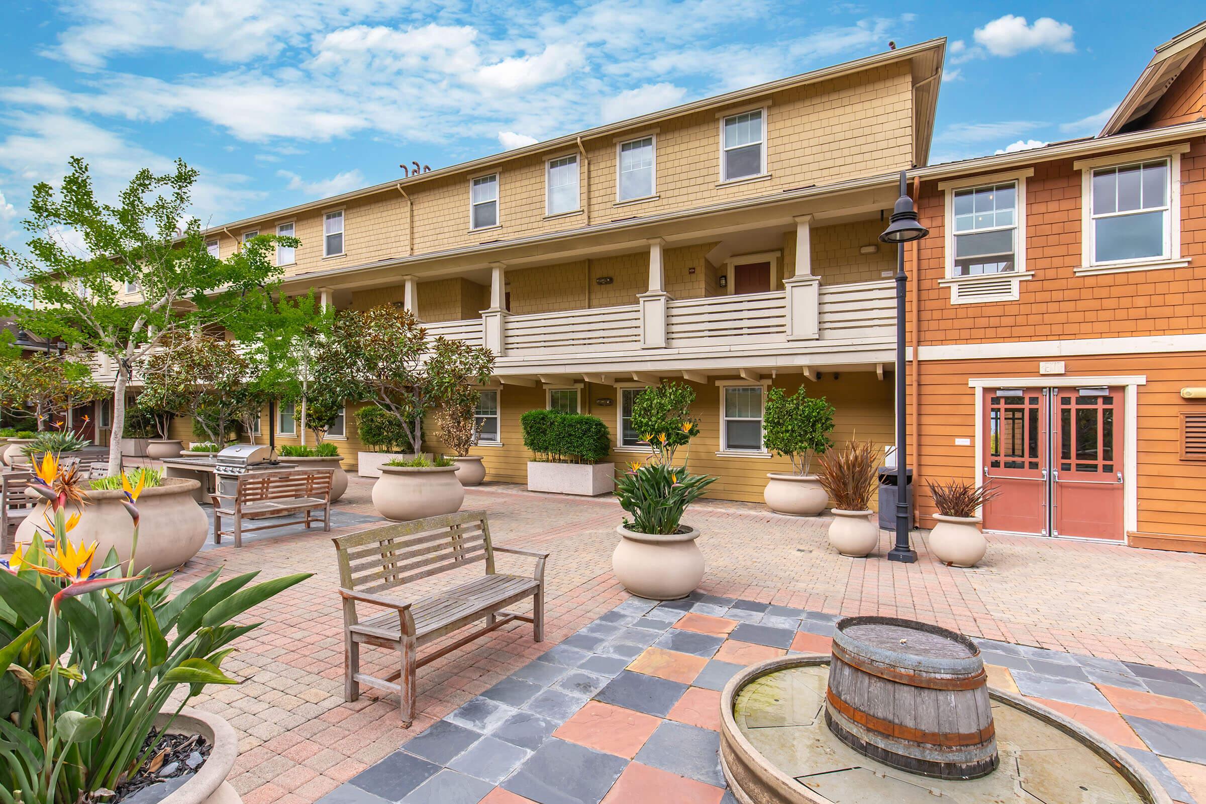 A spacious courtyard featuring potted plants and seating areas in front of two multi-story buildings with a warm color palette. The scene is bright and inviting, with greenery and decorative elements adding to the ambiance. A wooden barrel and colorful pavement tiles contribute to the outdoor aesthetic.