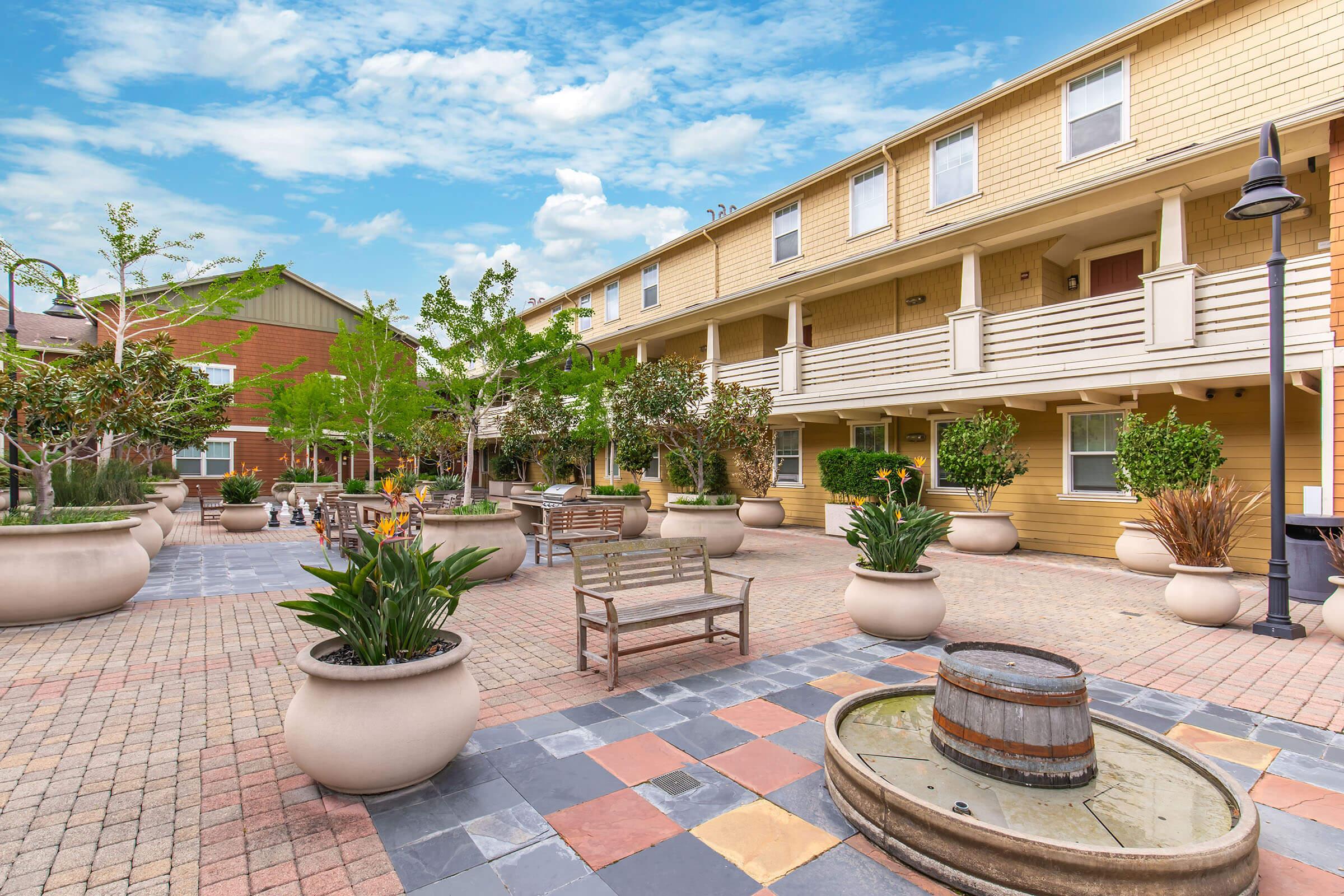 A well-maintained courtyard featuring a variety of large planters with greenery, wooden benches, and a decorative water feature. The background includes multi-story buildings with a bright blue sky above, creating a welcoming outdoor space for relaxation and social gatherings.