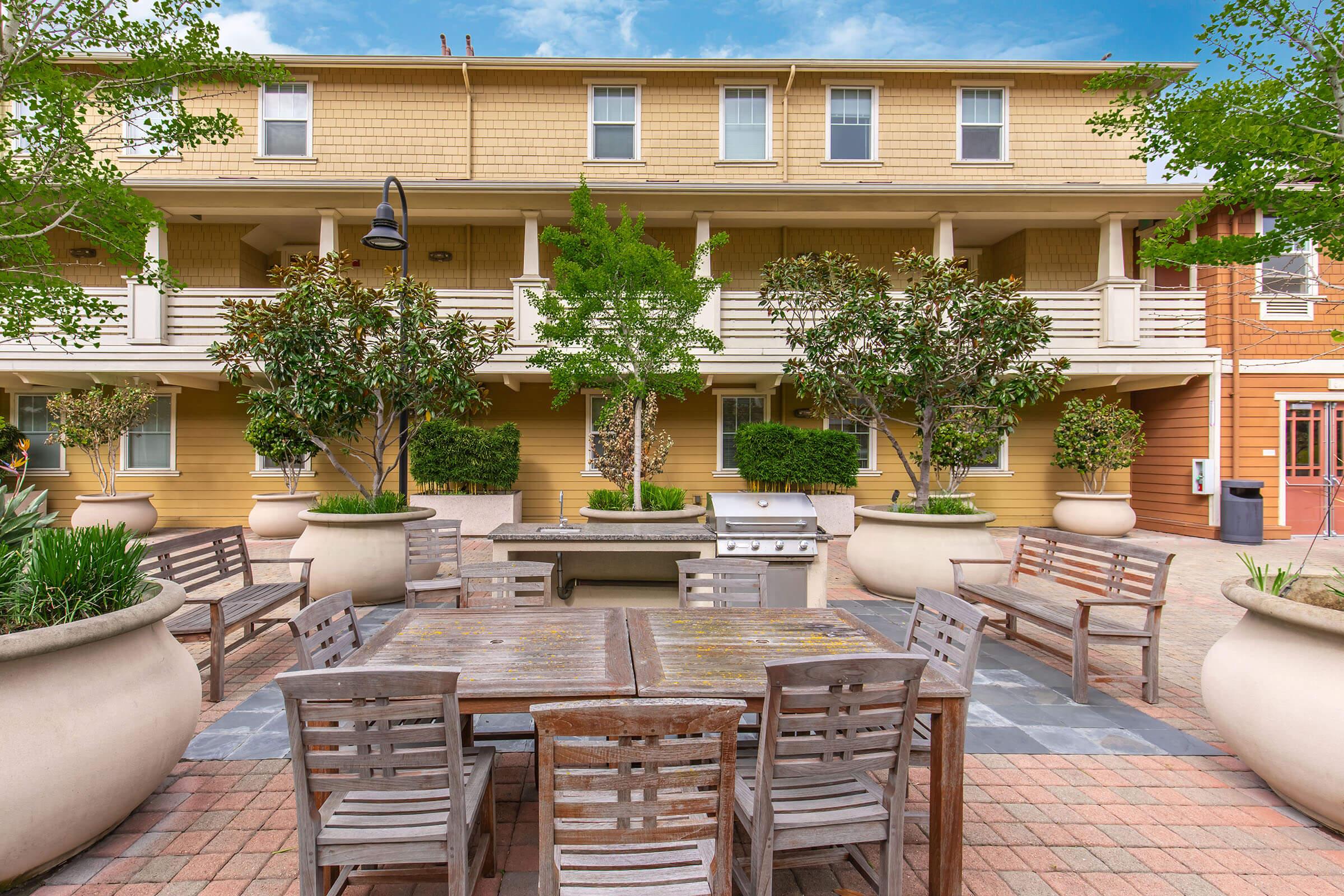 A well-maintained outdoor courtyard featuring wooden furniture, large planters with greenery, and a cooking grill area. The background shows a multi-story building with balconies, creating a welcoming and communal atmosphere. The sky is clear and blue, adding to the inviting setting.