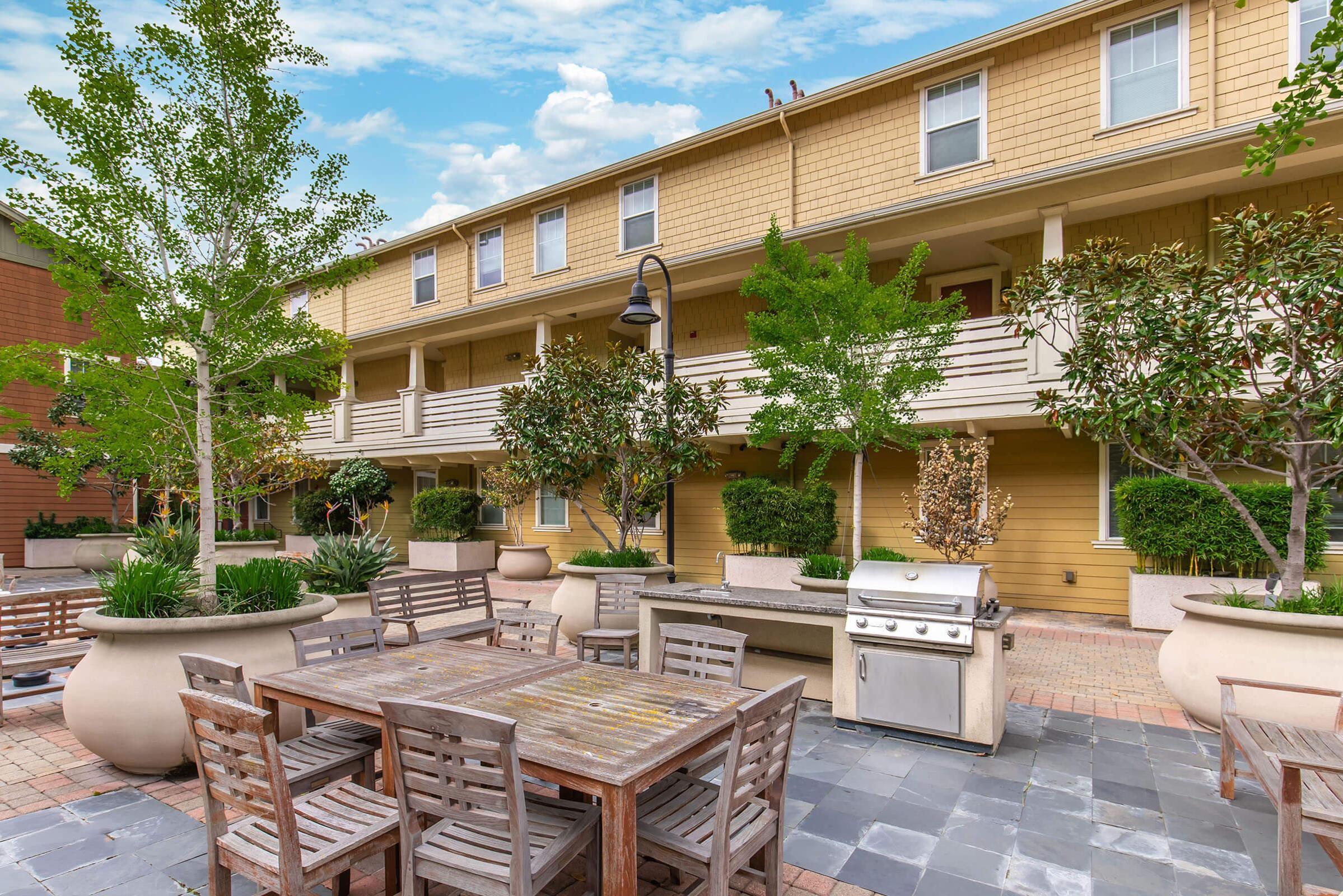 Outdoor courtyard featuring a patio area with a wooden dining table and chairs, potted trees, and decorative plants. In the background, a two-story building with a yellow exterior and balconies is visible. A barbecue grill is situated near the dining area on a stone-paved surface.