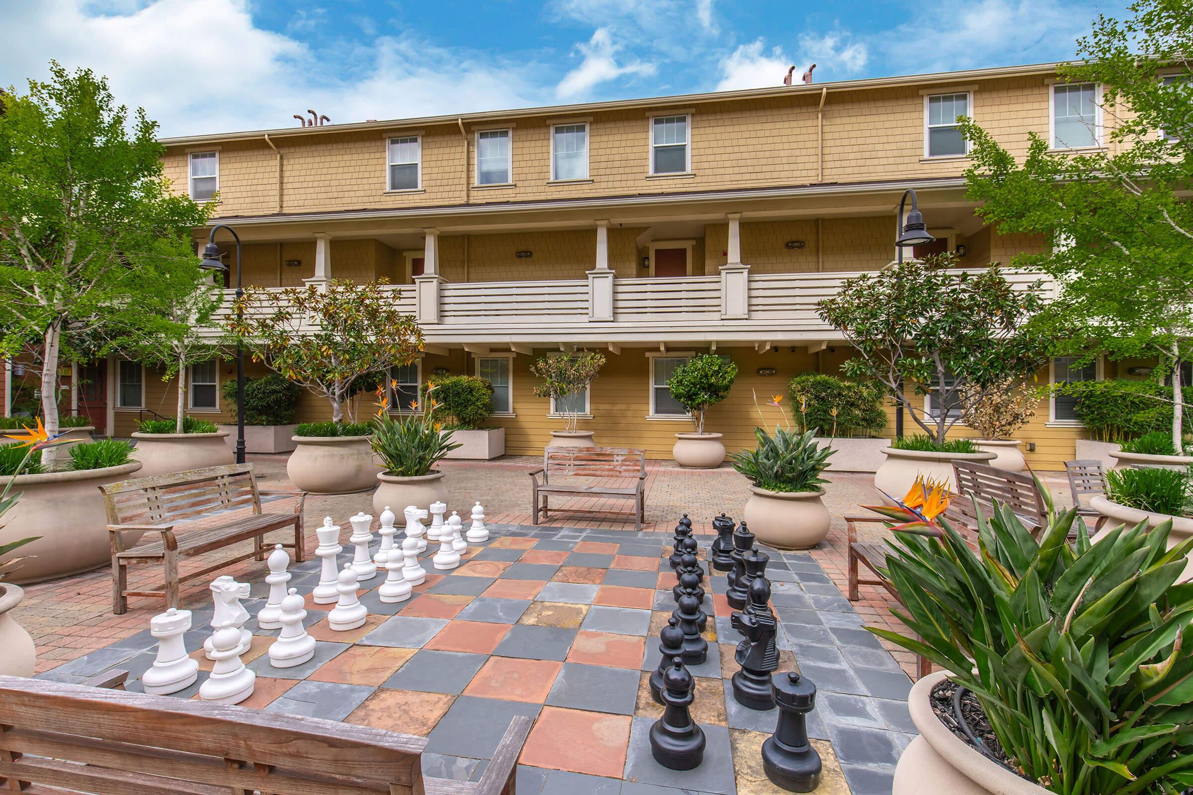 Outdoor area featuring a large chessboard with oversized white and black chess pieces. Surrounding the chessboard are benches and planters with lush greenery. In the background, there are modern buildings with a warm color palette and balconies. The sky is partly cloudy, creating a pleasant atmosphere.