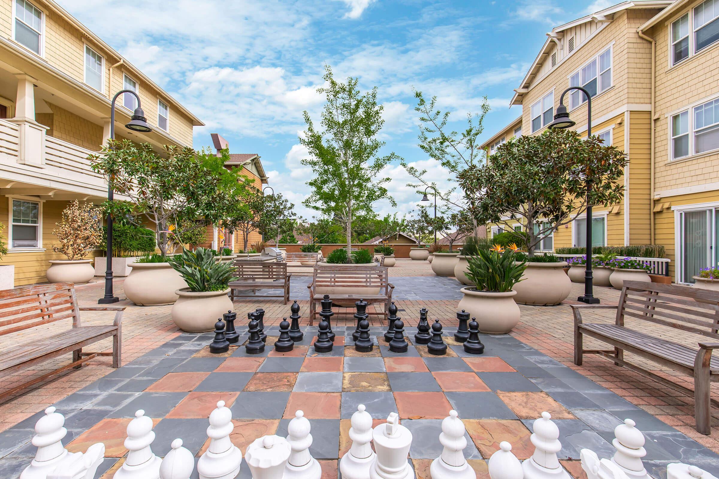 An outdoor courtyard featuring a large chessboard set between two sets of black and white chess pieces. Surrounded by potted plants and trees, the area includes several wooden benches and looks inviting with a clear blue sky overhead. Residential buildings are visible in the background.