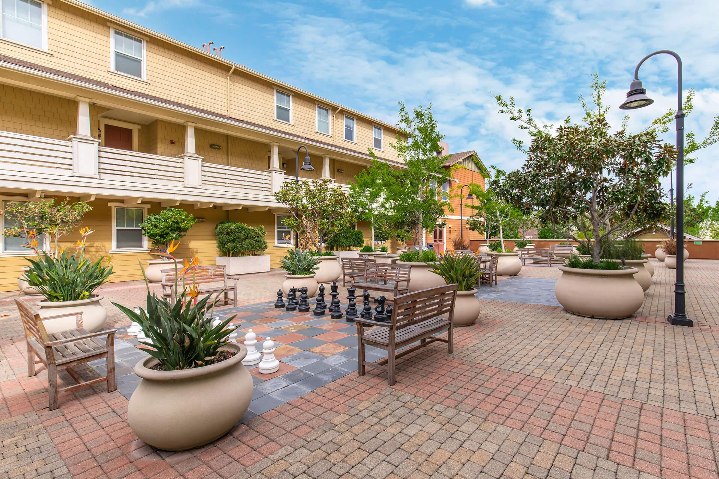 A spacious outdoor courtyard featuring several large planters with greenery, benches for seating, and a large chess set. Surrounding buildings in a warm color palette are visible in the background under a clear blue sky. The area is well-maintained and inviting, ideal for relaxation and leisure activities.