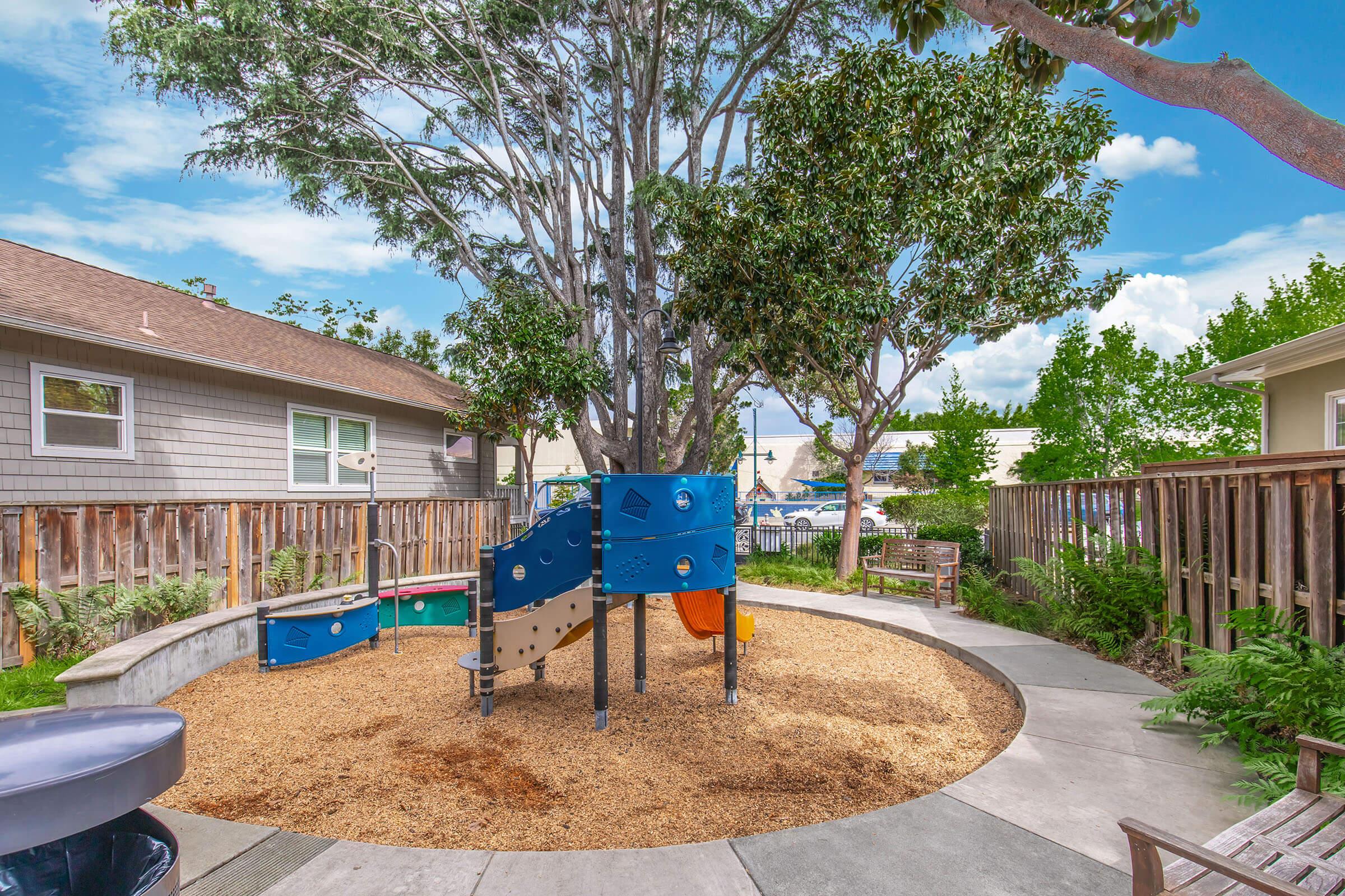 A small playground featuring colorful climbing structures surrounded by gravel and wood fencing. There are benches for seating, greenery with trees, and a clear blue sky in the background. The area is designed for children’s play and is located in a residential setting.