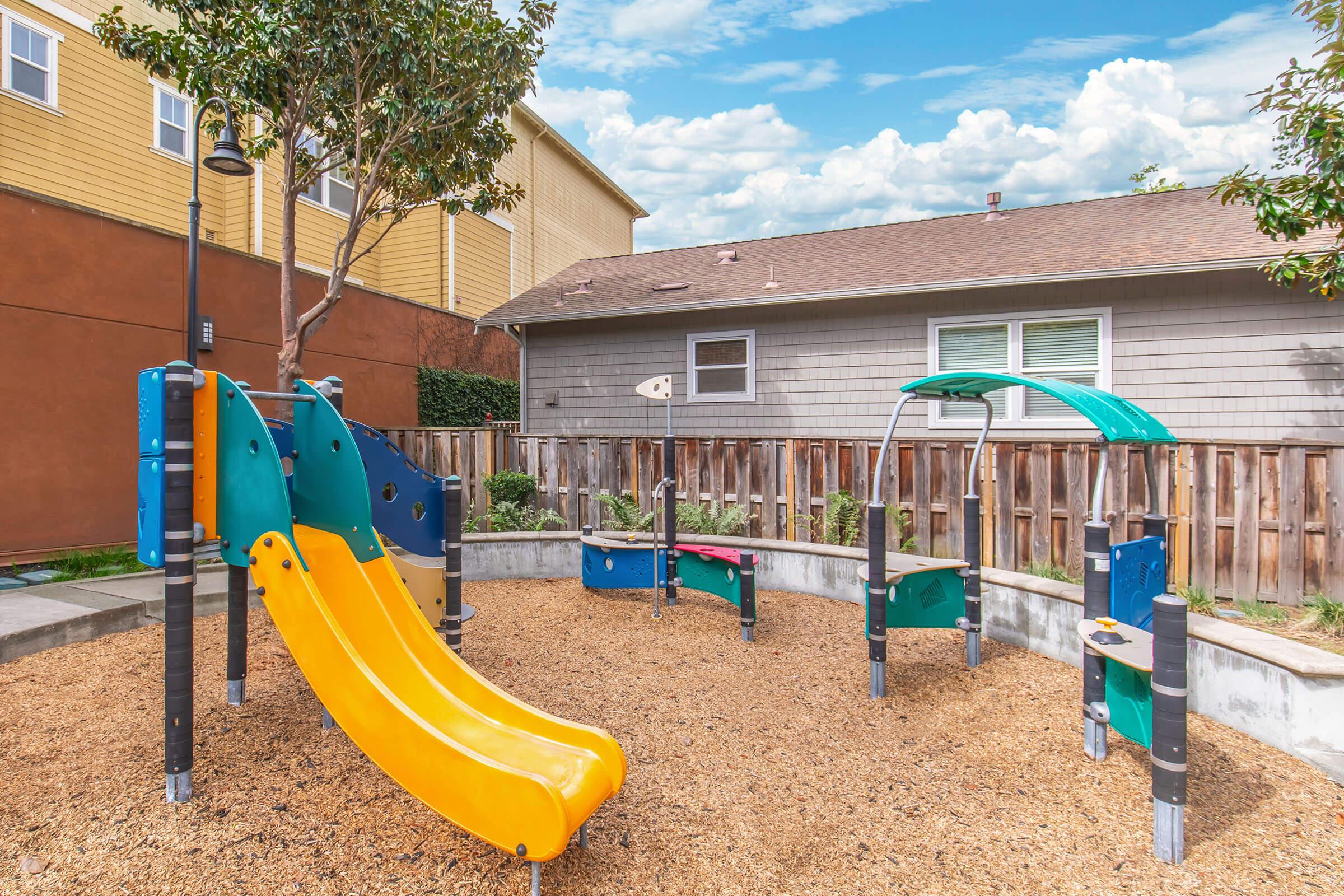 Colorful playground equipment featuring yellow and green slides, climbing structures, and swings, situated on a bed of wood chips. The playground is surrounded by wooden fencing and nearby residential buildings under a partly cloudy sky.