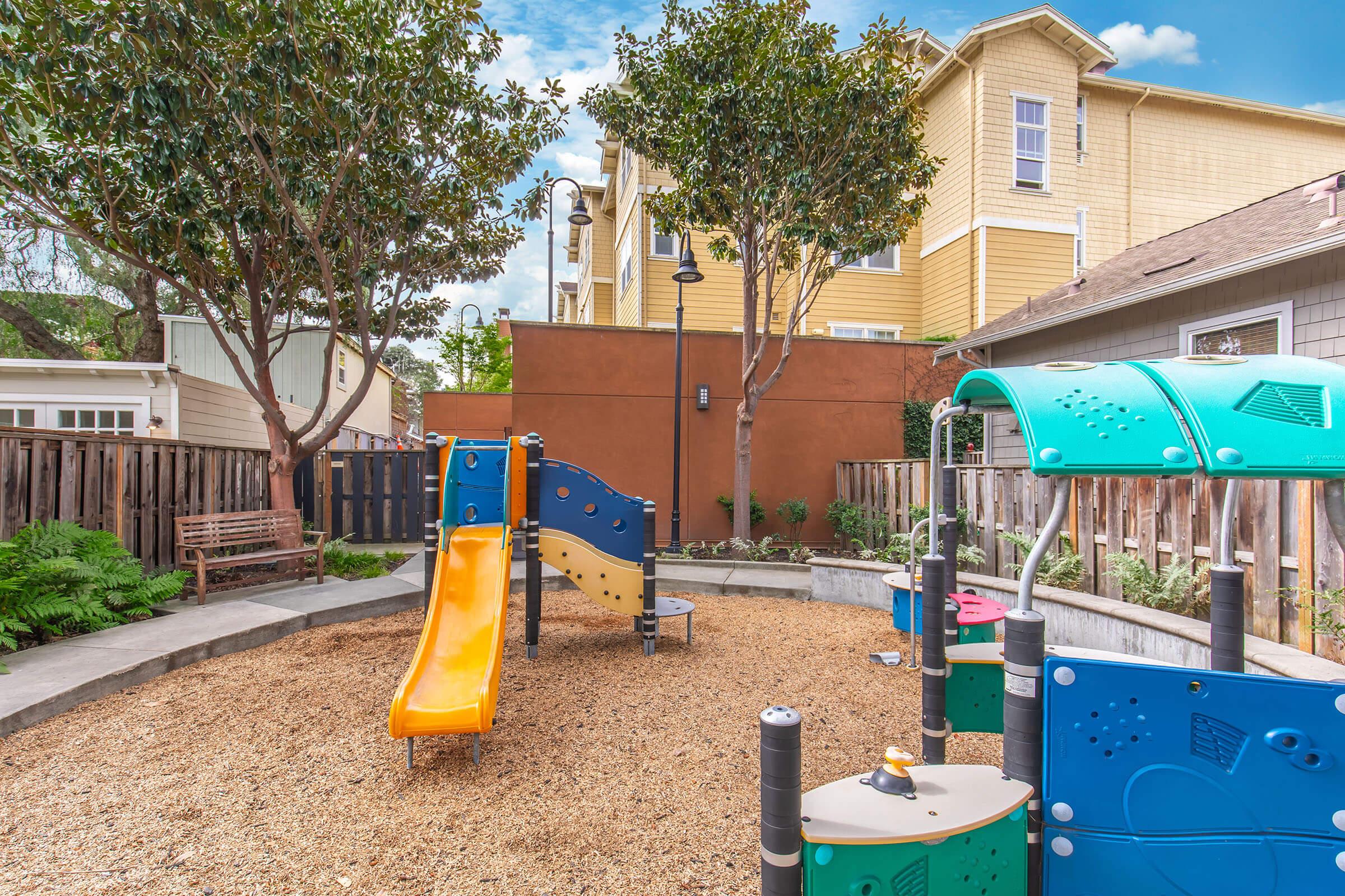 Colorful playground equipment with a yellow slide and climbing structure, surrounded by gravel and greenery. A wooden bench is visible in the background, along with a wall painted brown and residential buildings. The scene is bright and inviting, suggesting a space for children to play.