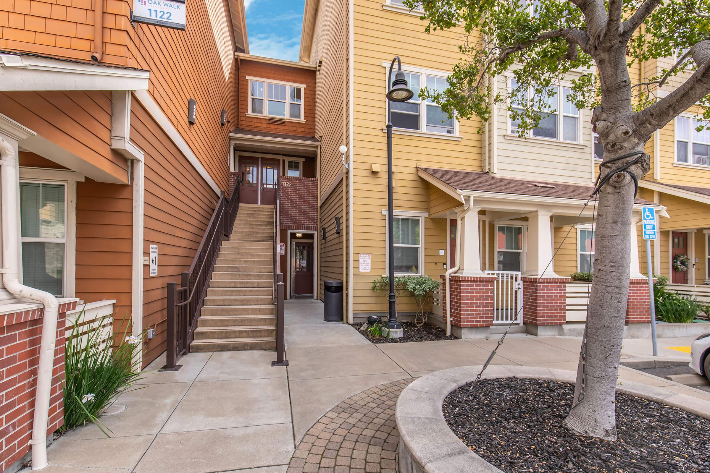Exterior view of a residential building featuring multiple units. The image shows a stairway leading up to a second floor, flanked by two colorful sections of the building—one orange and one yellow. A small tree and landscaped area are visible in the foreground, along with a lamp post and a wheelchair-accessible sign.