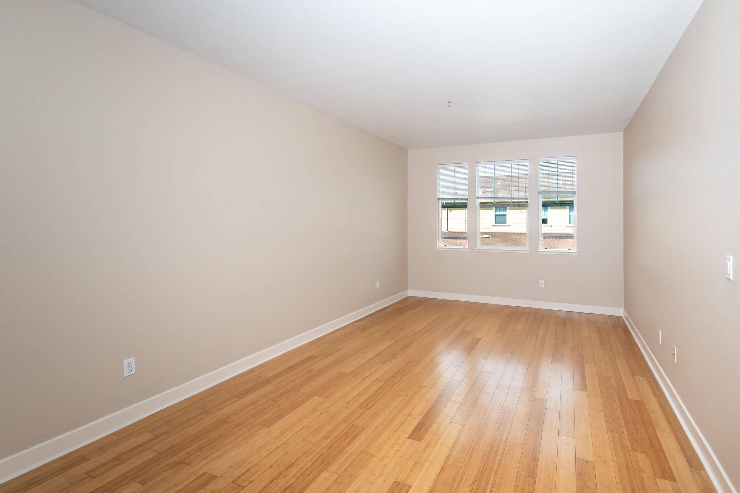 A spacious, empty room with light-colored walls and bamboo flooring. Three large windows allow natural light to fill the space, offering a view of the outdoors. The room has a clean, minimalist aesthetic, perfect for various layout options.
