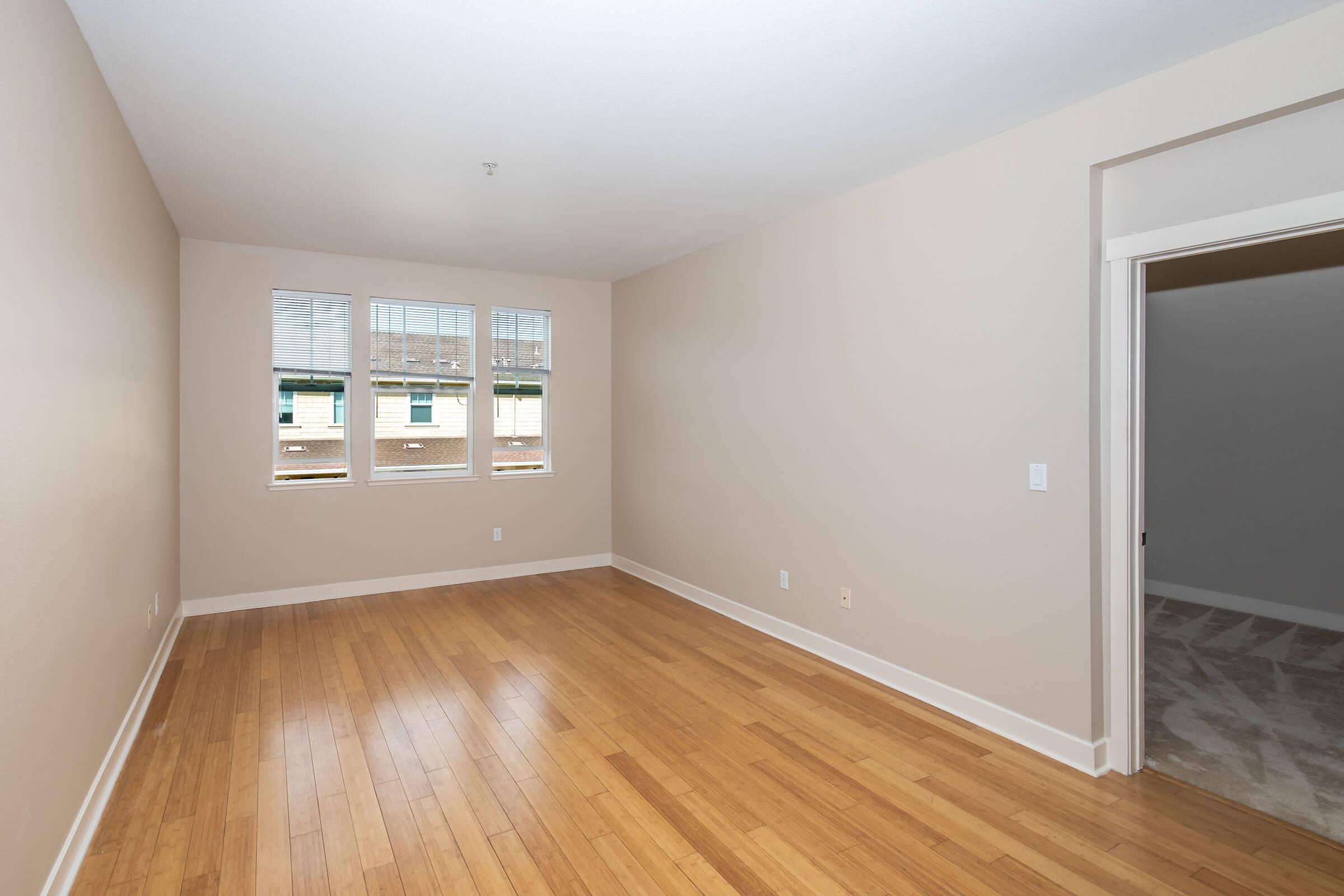 A spacious, empty room with light beige walls and hardwood flooring. There are three windows allowing natural light, and a doorway leading to another space. The area is uncluttered, creating a minimalist, airy feel.