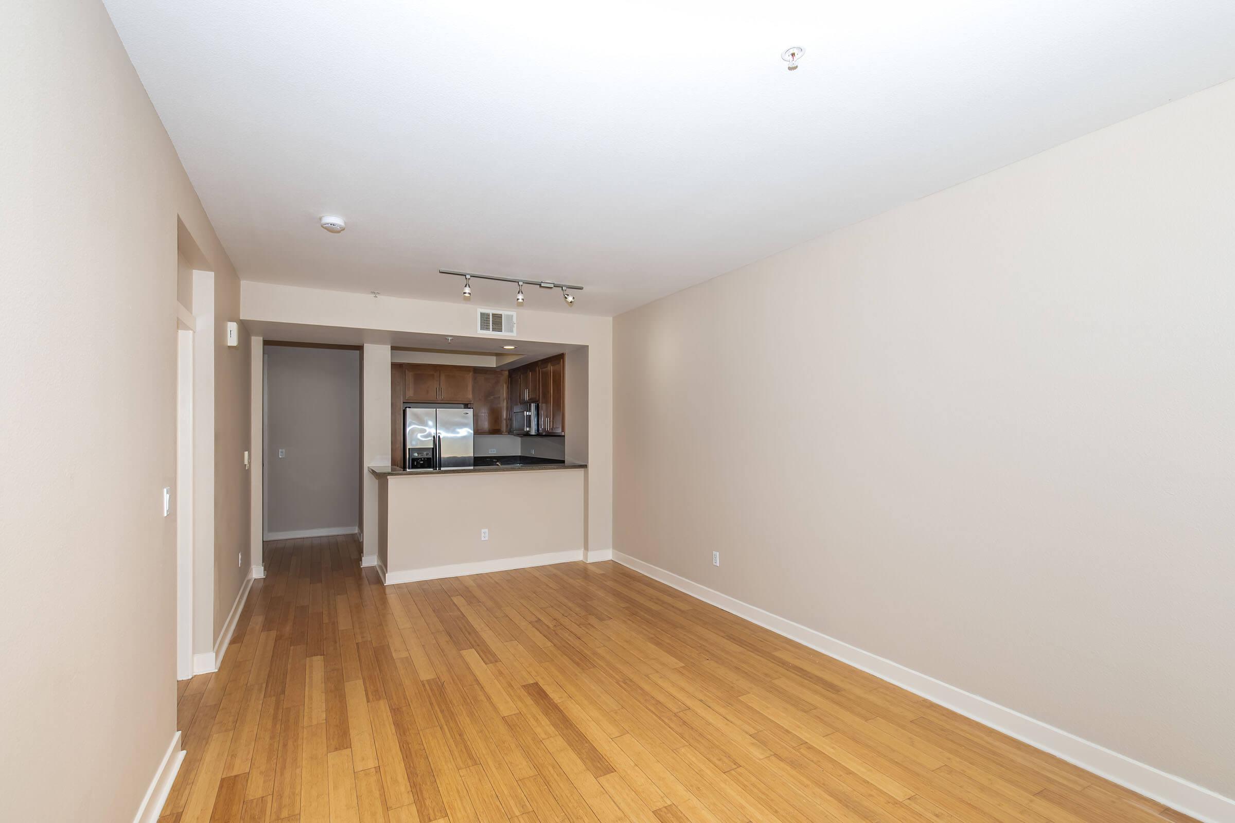Spacious, empty living area featuring light-colored walls and bamboo flooring. The room has a doorway leading to a kitchen area, which includes wooden cabinetry and stainless steel appliances. Natural light fills the space, creating a bright and airy atmosphere. Suitable for various furniture arrangements.