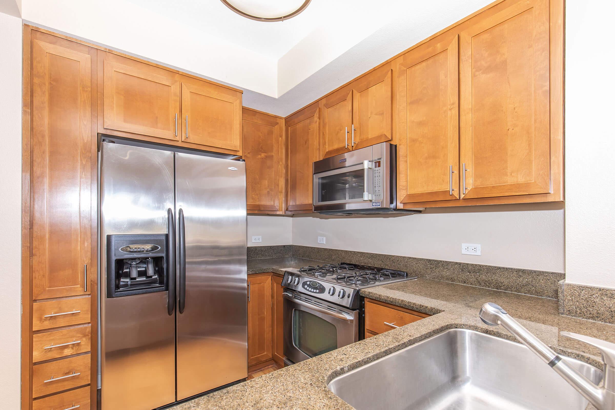 A modern kitchen featuring light wooden cabinetry, stainless steel appliances including a fridge, oven, and microwave. The countertop is made of granite, and there's a double sink with a faucet. The layout is compact and functional, with a bright atmosphere created by the overhead lighting.