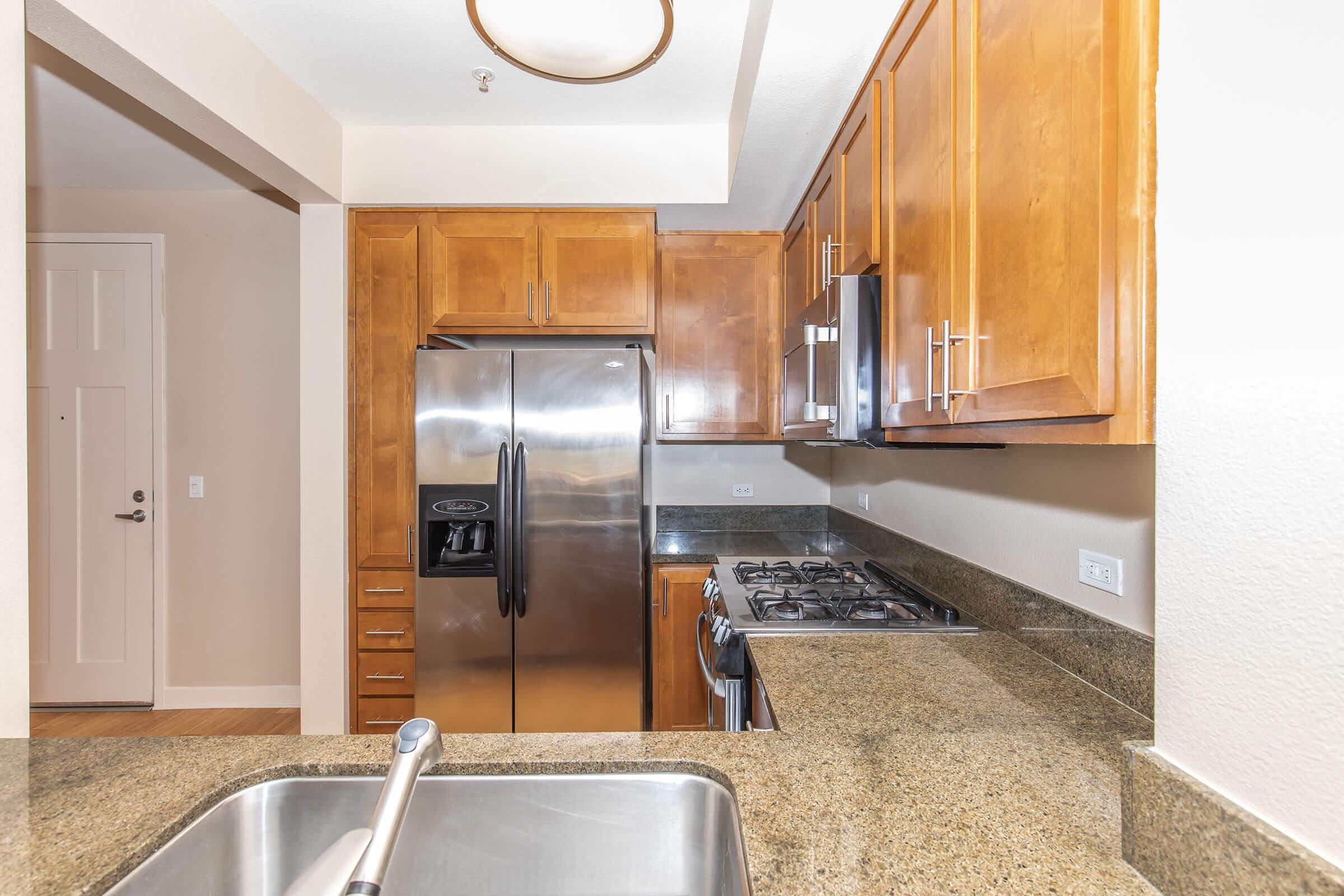 A modern kitchen with wooden cabinets, stainless steel refrigerator, and gas stove. The countertop is made of granite, and there is a sink with a faucet. The walls are painted in a light color, and there is a door leading to an entrance area in the background. Bright and inviting interior space.