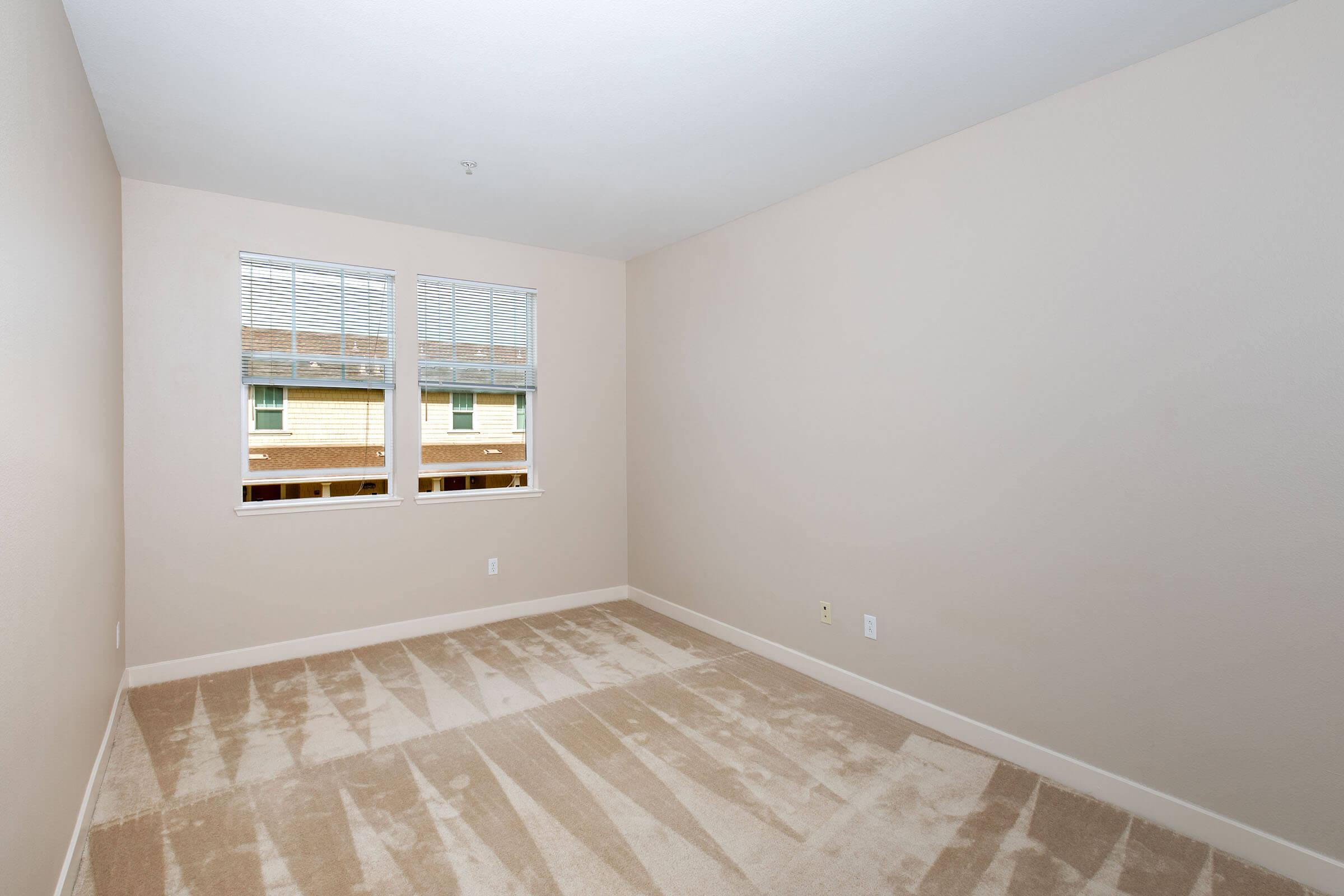 Empty bedroom with beige walls and carpet. Two windows let in natural light, revealing an outdoor view. The space appears clean and uncluttered, ideal for personal decoration or furnishing.