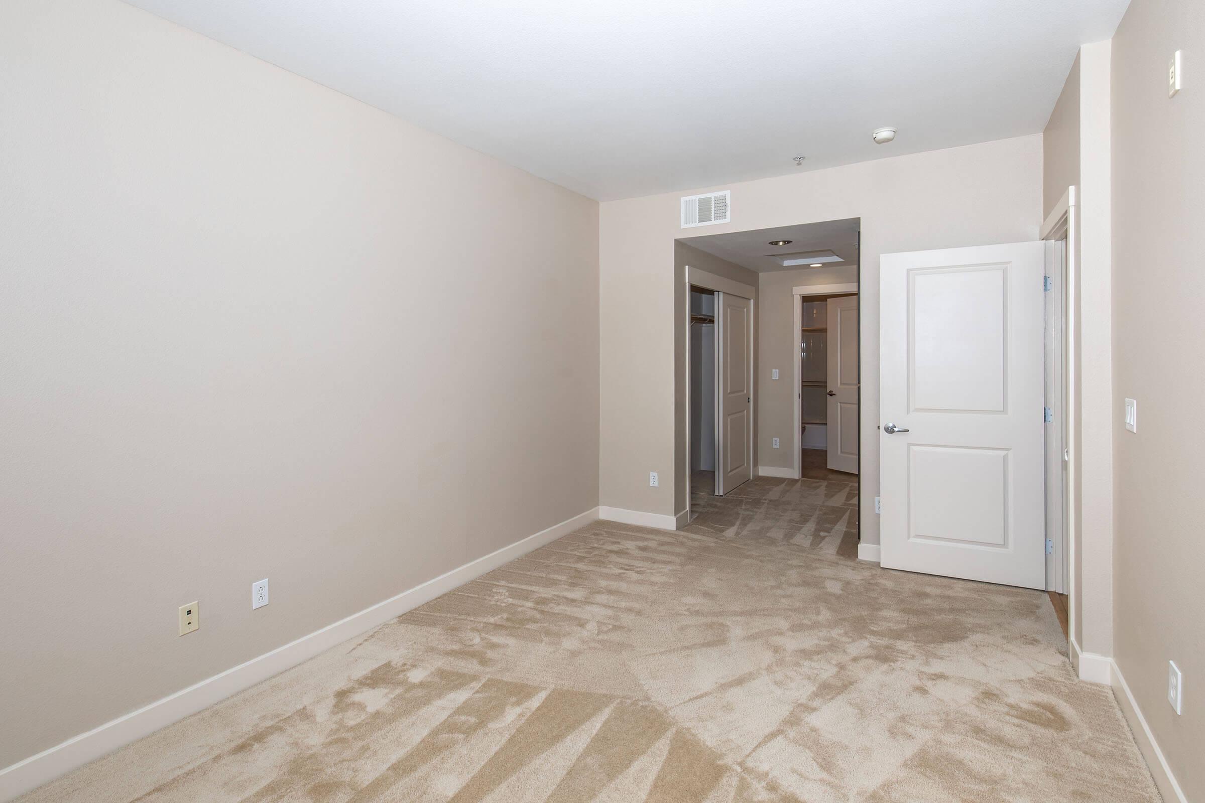 A vacant room with light beige walls and soft carpet. An open door leads to a hallway, and another door is visible on the left, suggesting access to a closet. The space is well-lit and designed for potential furnishings, with no visible decorations.