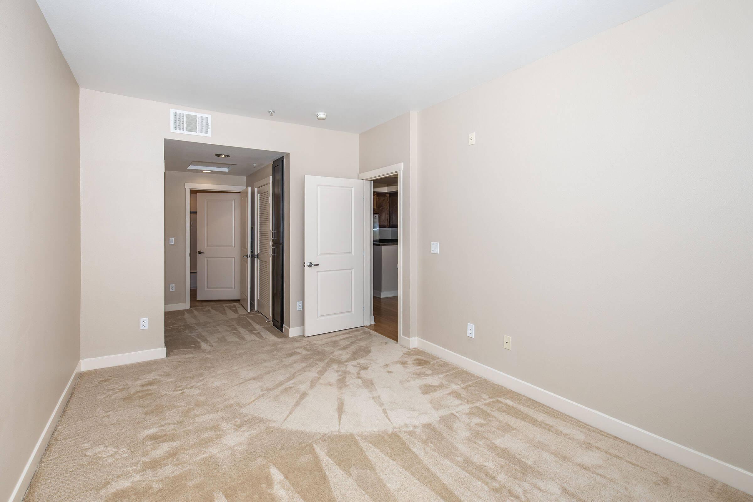 Empty room with beige walls and carpet. There are two doors: one leading to a closet and another to a hallway. The lighting is bright, and the space appears clean and uncluttered, suitable for various uses such as a bedroom or office.