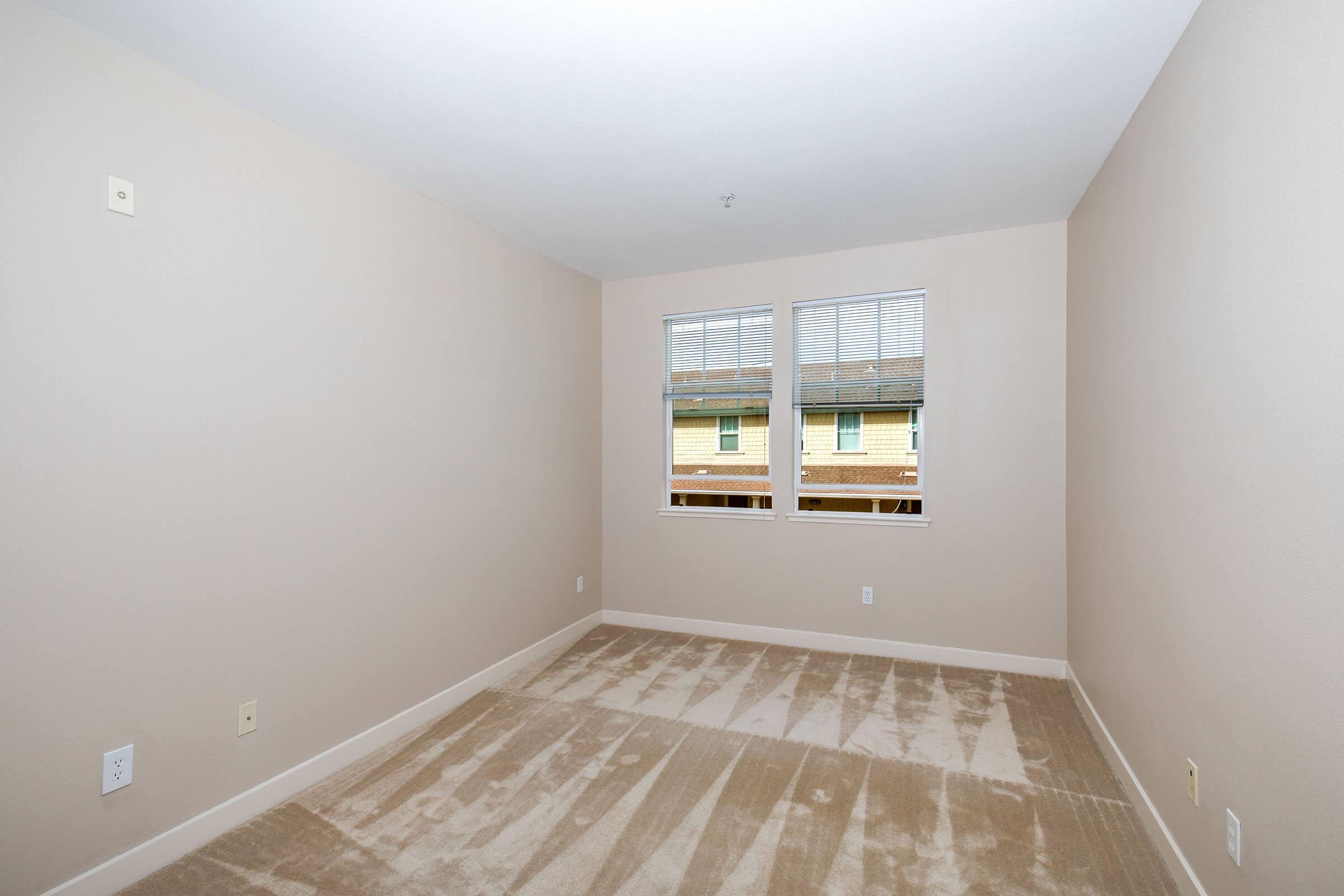 A vacant room with light beige walls and carpet. It features two windows with blinds, letting in natural light. The space is unfurnished, providing a blank canvas for personalization. The room appears clean and well-maintained, with minimal decor and no visible furniture.