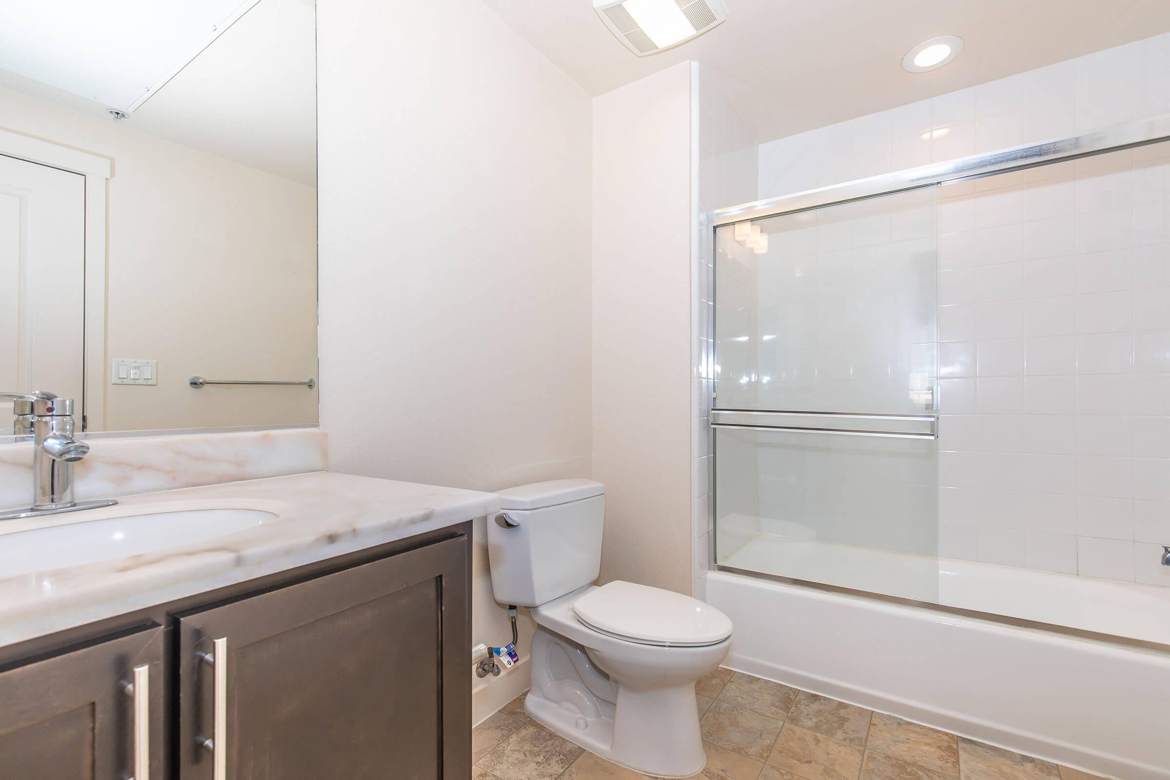 A clean and modern bathroom featuring a white shower-tub combination, a sleek mirrored vanity, a toilet, and light-colored tile flooring. The walls are painted in a neutral tone, enhancing the spacious feel of the room.