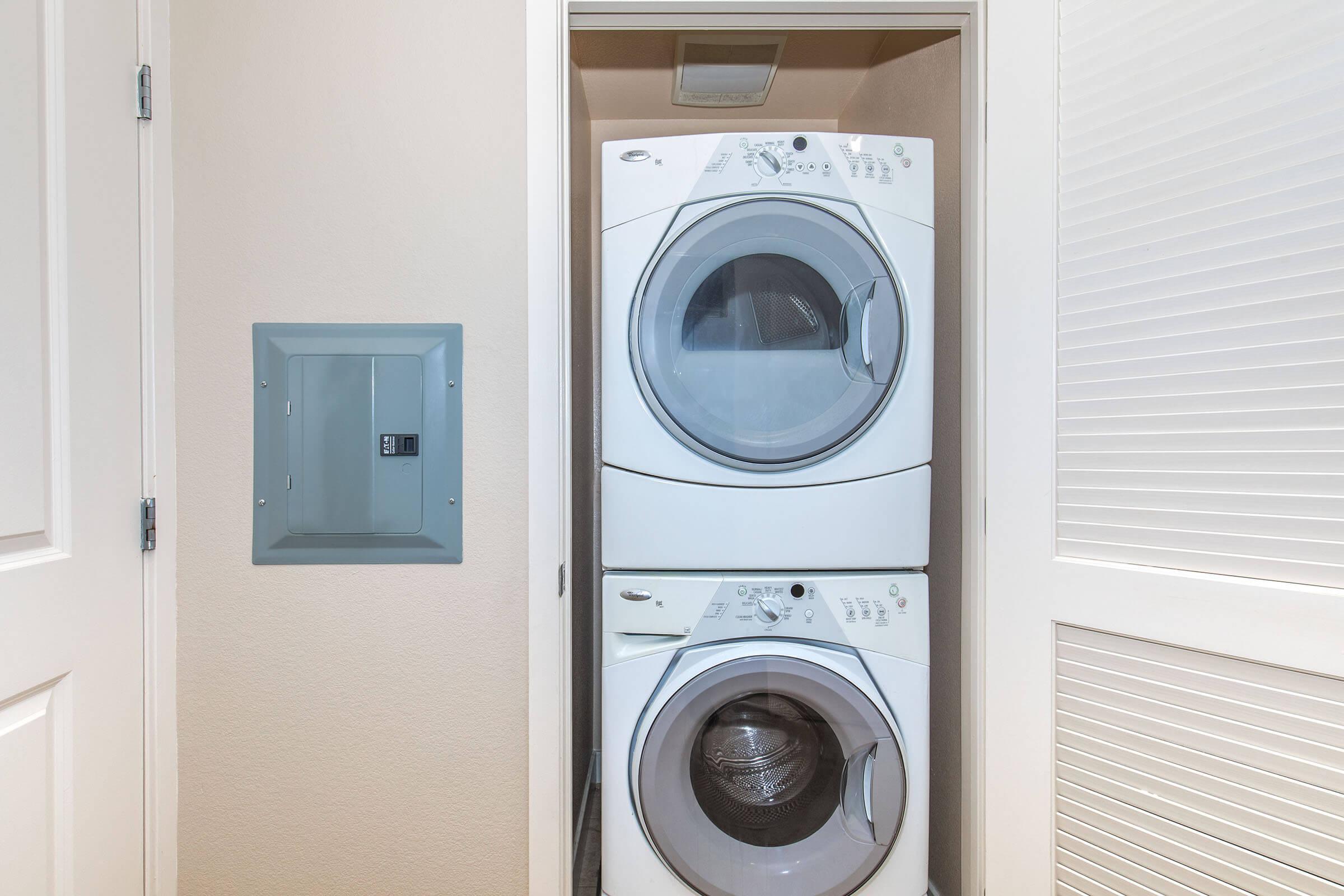 Stacked washer and dryer in a compact laundry space, with a gray electrical panel mounted on the wall next to them. The laundry appliances are white, and the surrounding walls are a neutral color, creating a clean and organized look. The door to the laundry area is slightly ajar.