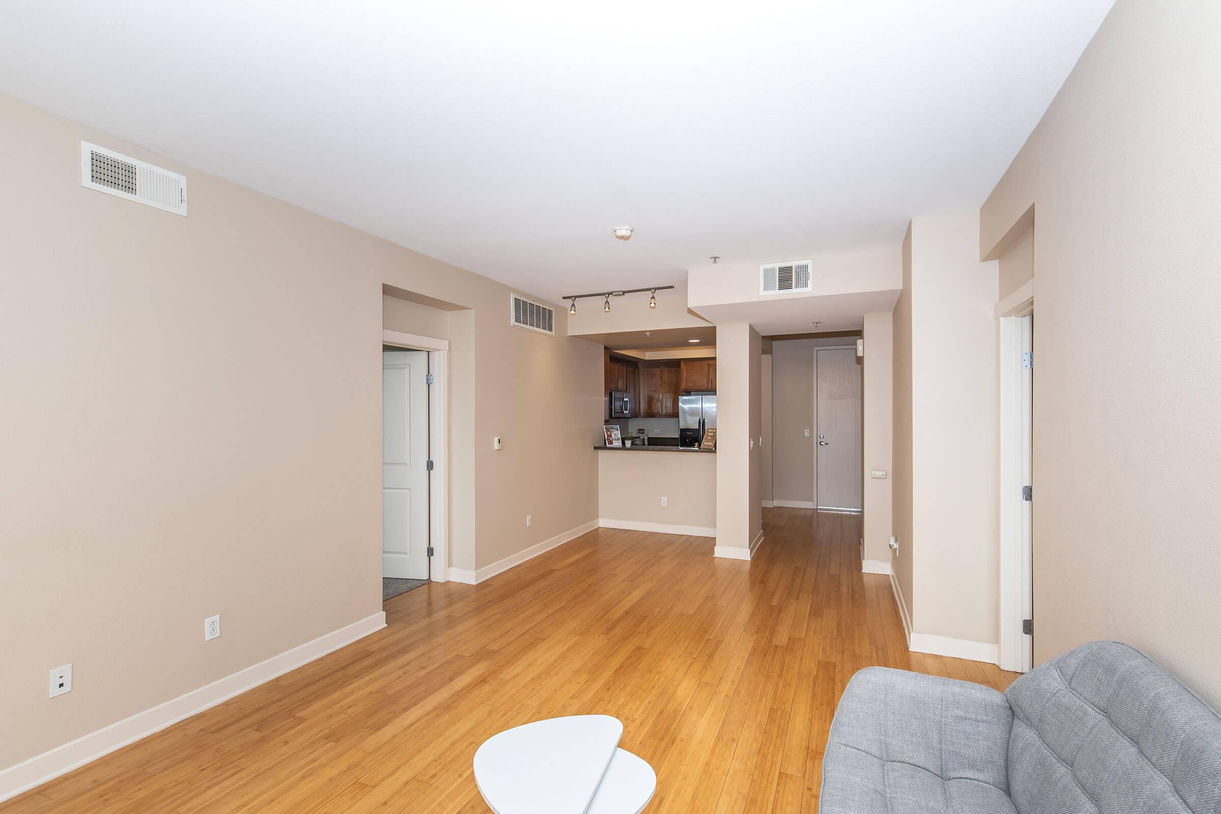 A spacious, empty living area featuring light brown walls and hardwood flooring. There’s a small round table in the foreground, and towards the back, a modern kitchen is visible. Doorways are seen on either side, leading to separate rooms, while natural light illuminates the space from the windows.