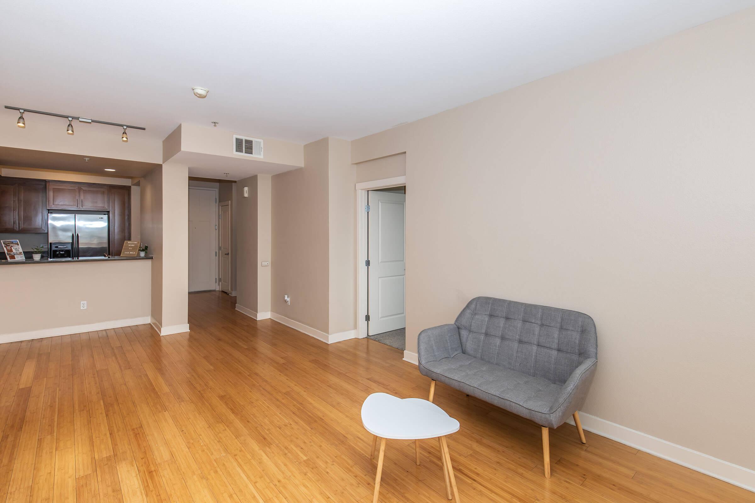 A spacious, modern living area featuring light beige walls and bamboo flooring. A gray upholstered armchair sits next to a small white table, while an open kitchen with dark wood cabinetry and bar seating is visible in the background. The room is well-lit and minimalist in design.