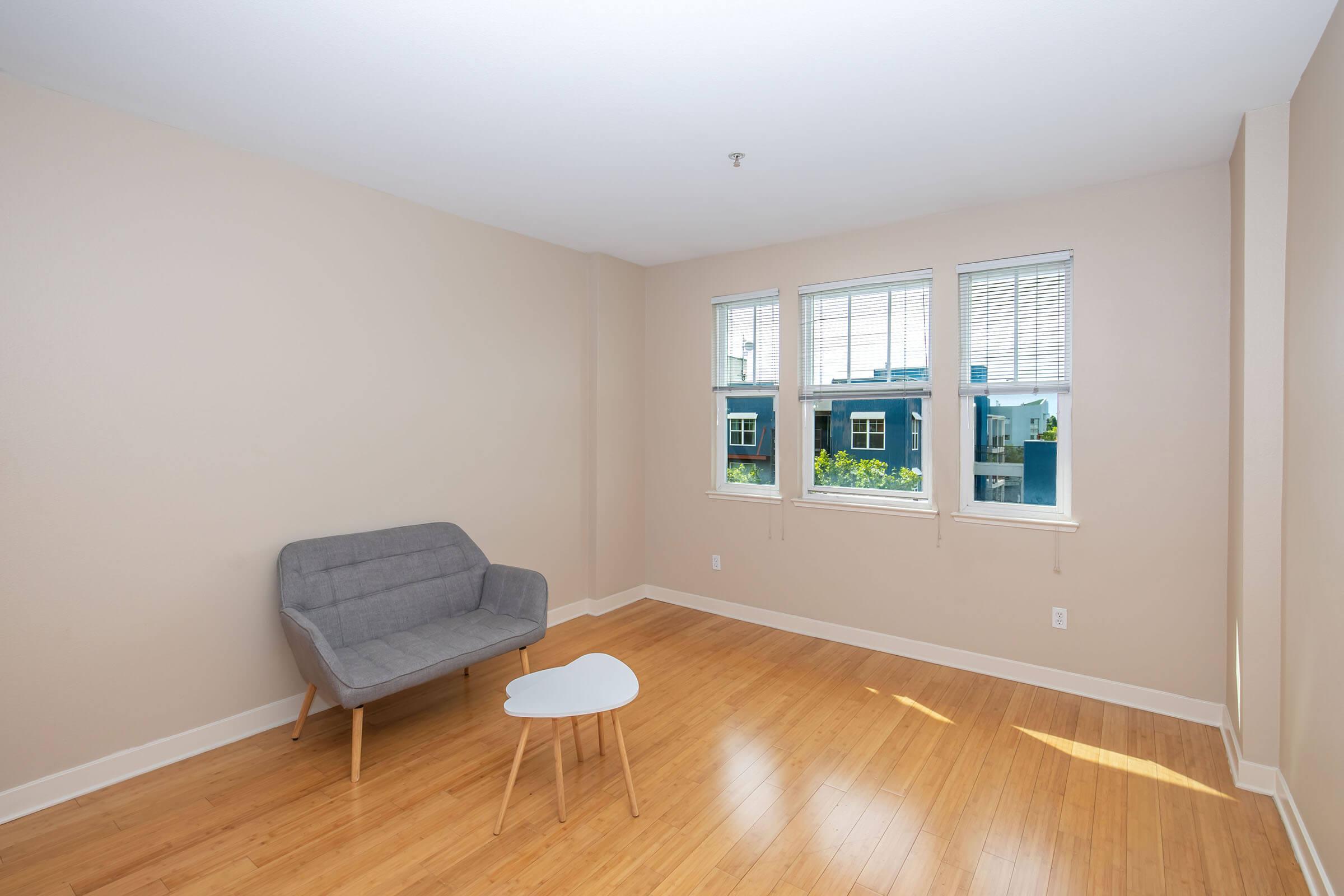 A bright, empty room with light beige walls and bamboo flooring. It features three windows allowing natural light, a gray sofa, and a small white coffee table. The view outside includes green trees.