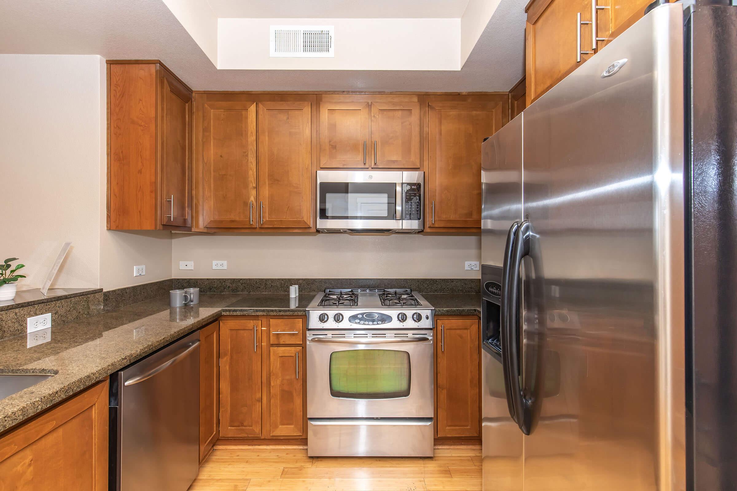 A modern kitchen featuring wooden cabinets, granite countertops, stainless steel appliances including a microwave, oven, and refrigerator. The kitchen has a clean, organized layout with hardwood flooring and a small plant on the counter. Natural light from a nearby window brightens the space.