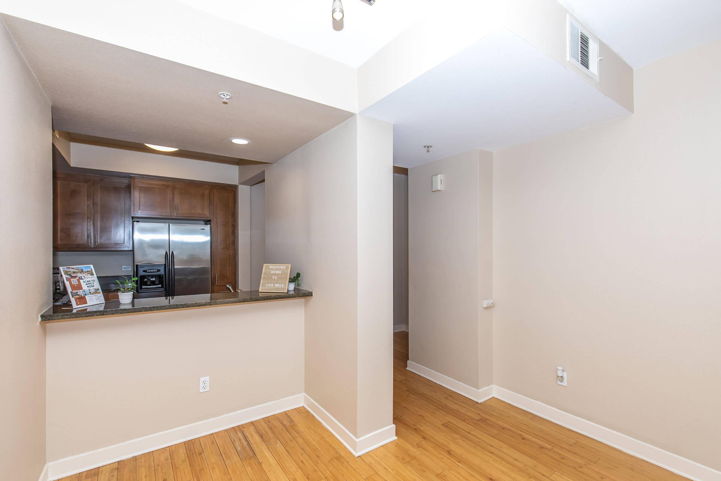 A view of a modern kitchen and dining area featuring a breakfast bar, stainless steel refrigerator, and wooden cabinets. The adjacent space has light-colored walls and hardwood flooring, creating an open and airy atmosphere. Natural light enhances the overall appeal of the room.