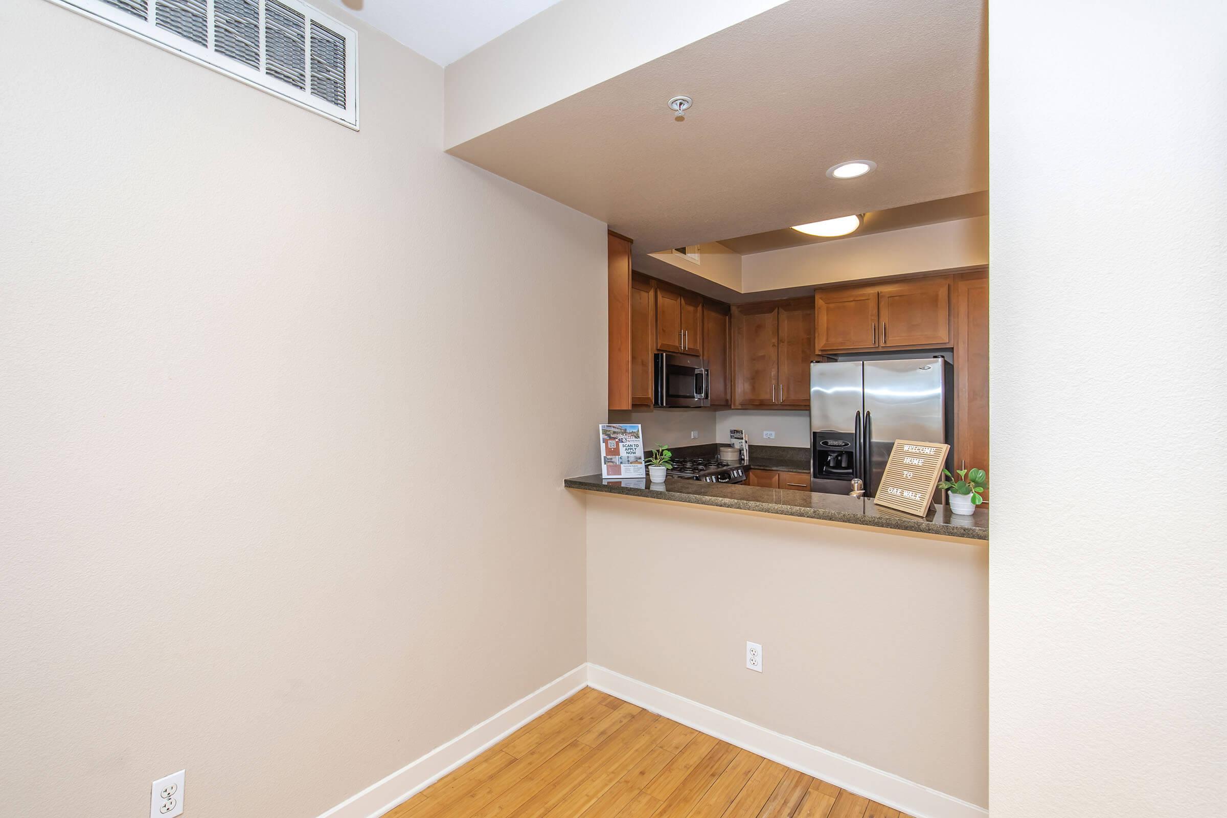 A kitchen area featuring wooden cabinets and stainless steel appliances, including a refrigerator and stove. The countertop is open to a small dining or living space. The walls are a light beige color, and there is natural wood flooring. A small plant and informational signs are placed on the countertop.