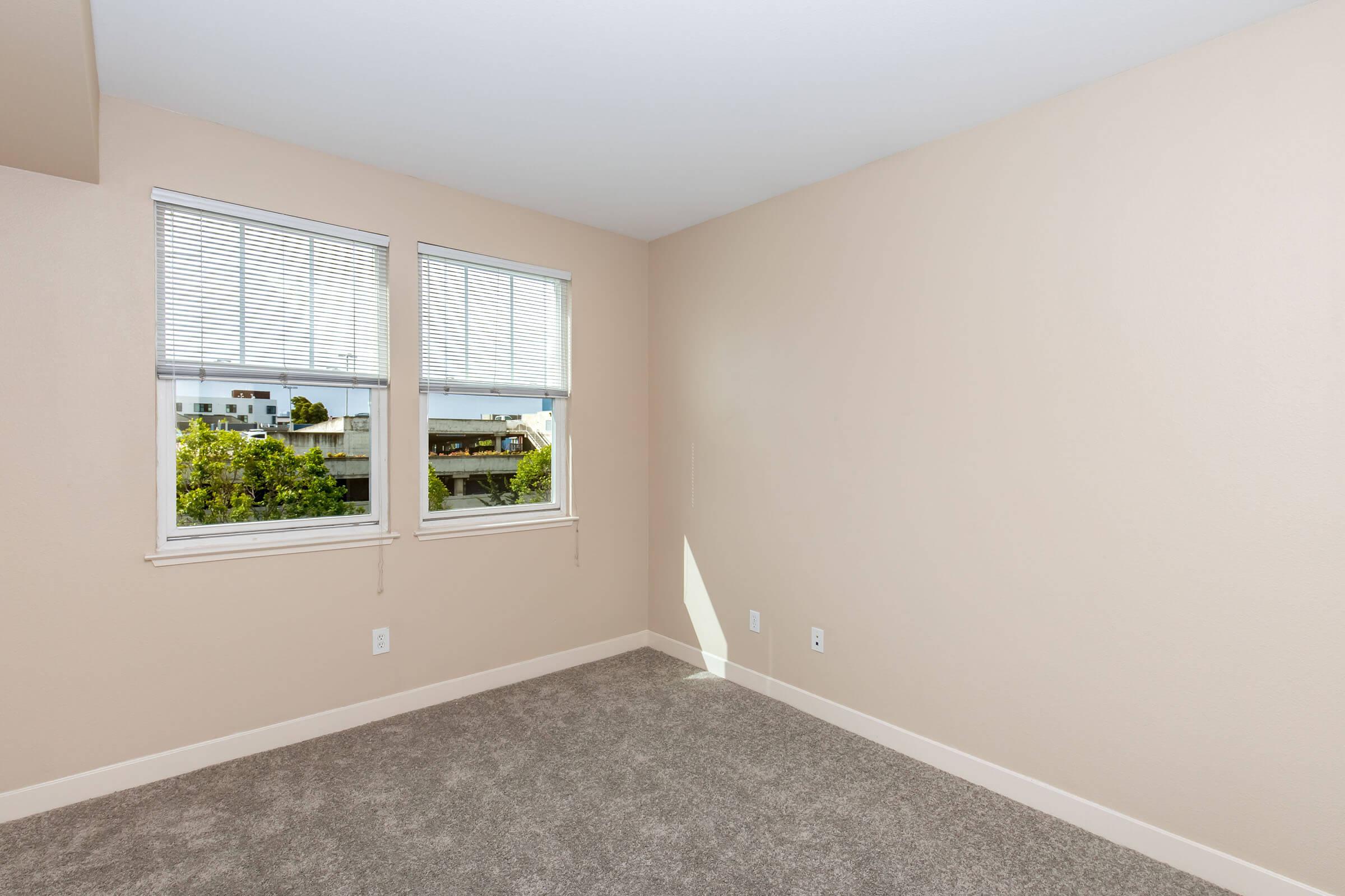 Empty room featuring light beige walls and plush gray carpeting. Two windows with white blinds allow natural light, offering a view of greenery outside. The space is unfurnished and has a clean, minimalist appearance.