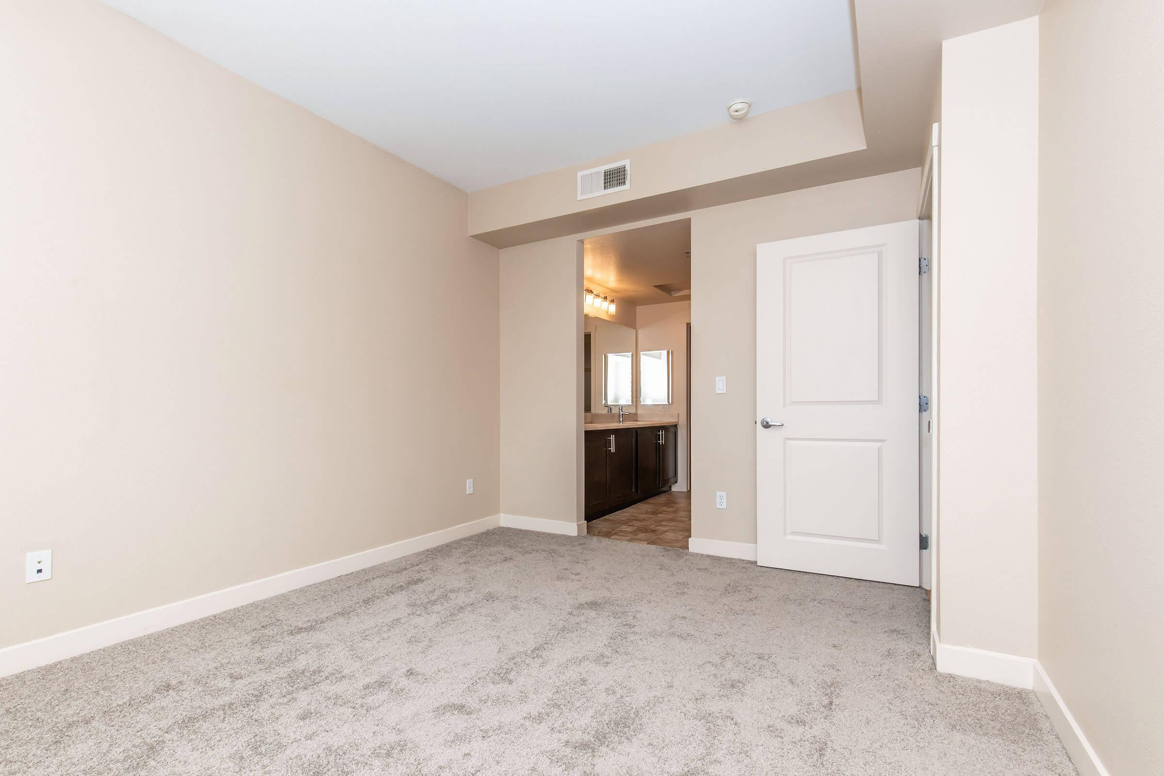 A vacant room with light beige walls and gray carpet flooring. The space features a doorway leading to a bathroom area, which includes a large mirror and vanity. Natural light illuminates the room, creating a bright and airy atmosphere.
