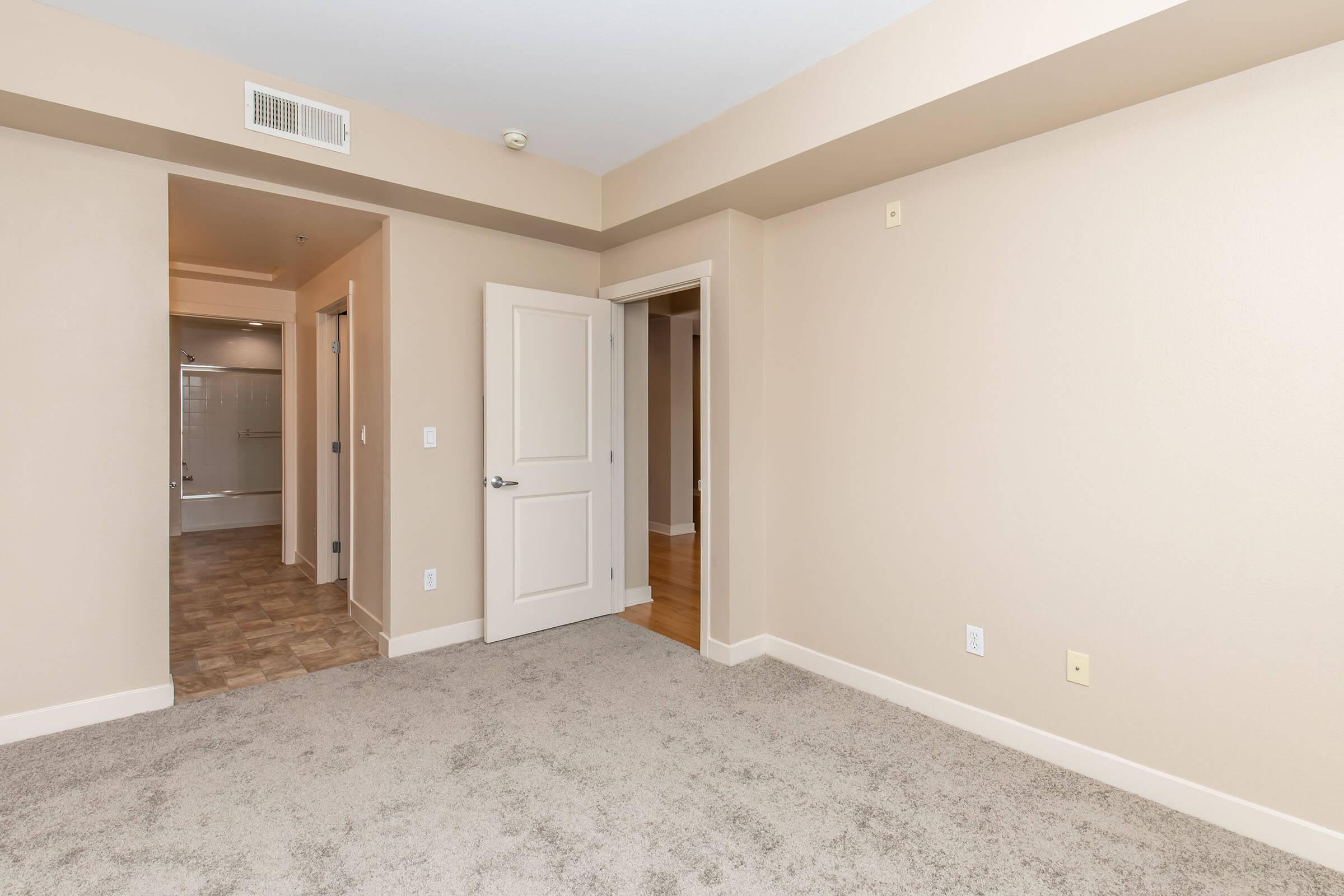Empty room with light beige walls and soft carpet flooring. The space features an open doorway leading to a hallway, with another door visible on the left. There is a visible bathroom area at the end of the hall, with a light tile pattern. Natural light is not prominent in this indoor setting.