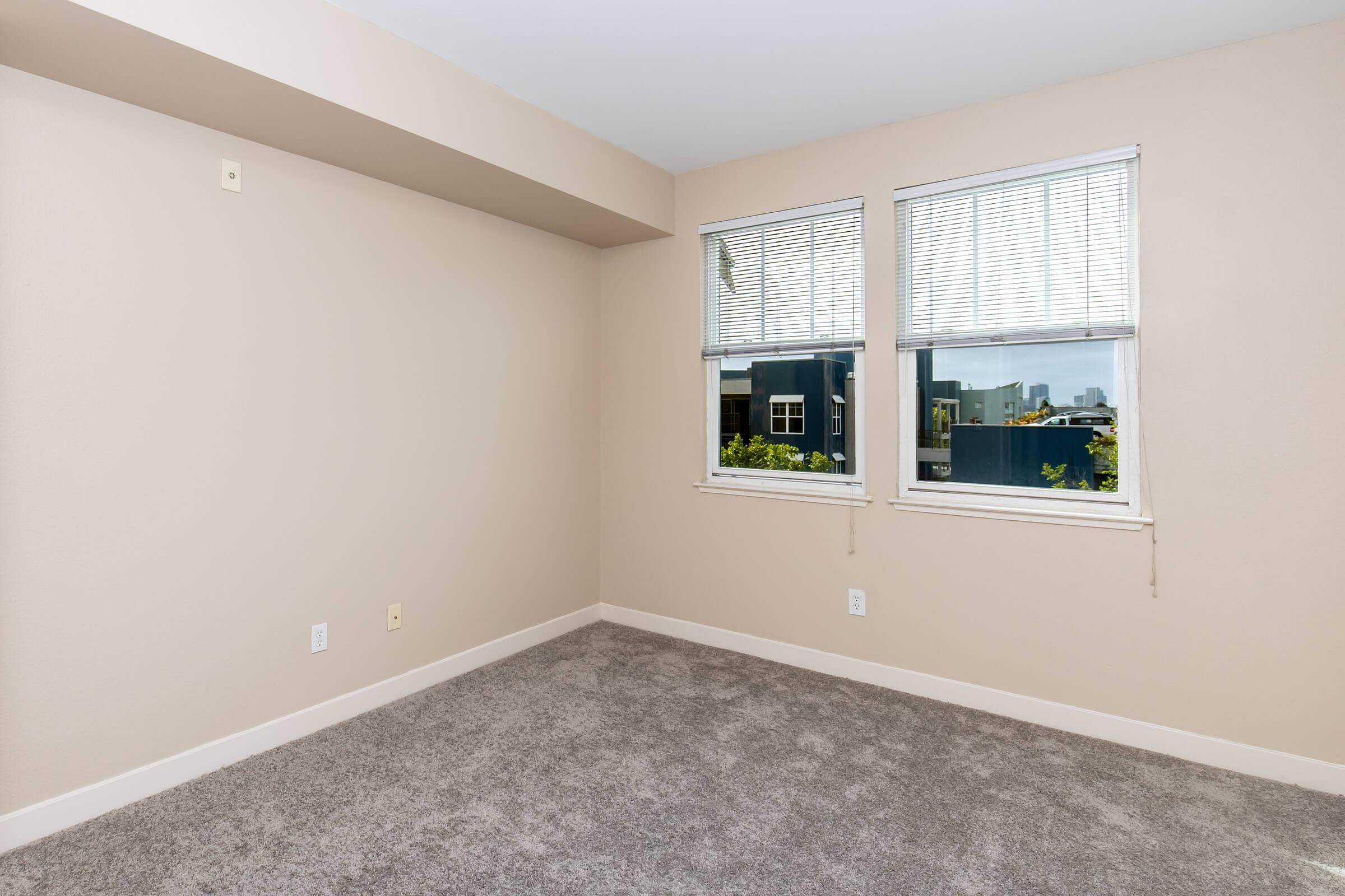 A corner of a room with light beige walls and gray carpet. There are two white-framed windows with mini blinds letting in natural light. The room appears empty, with no furniture or decor, and offers a view of nearby buildings through the windows.