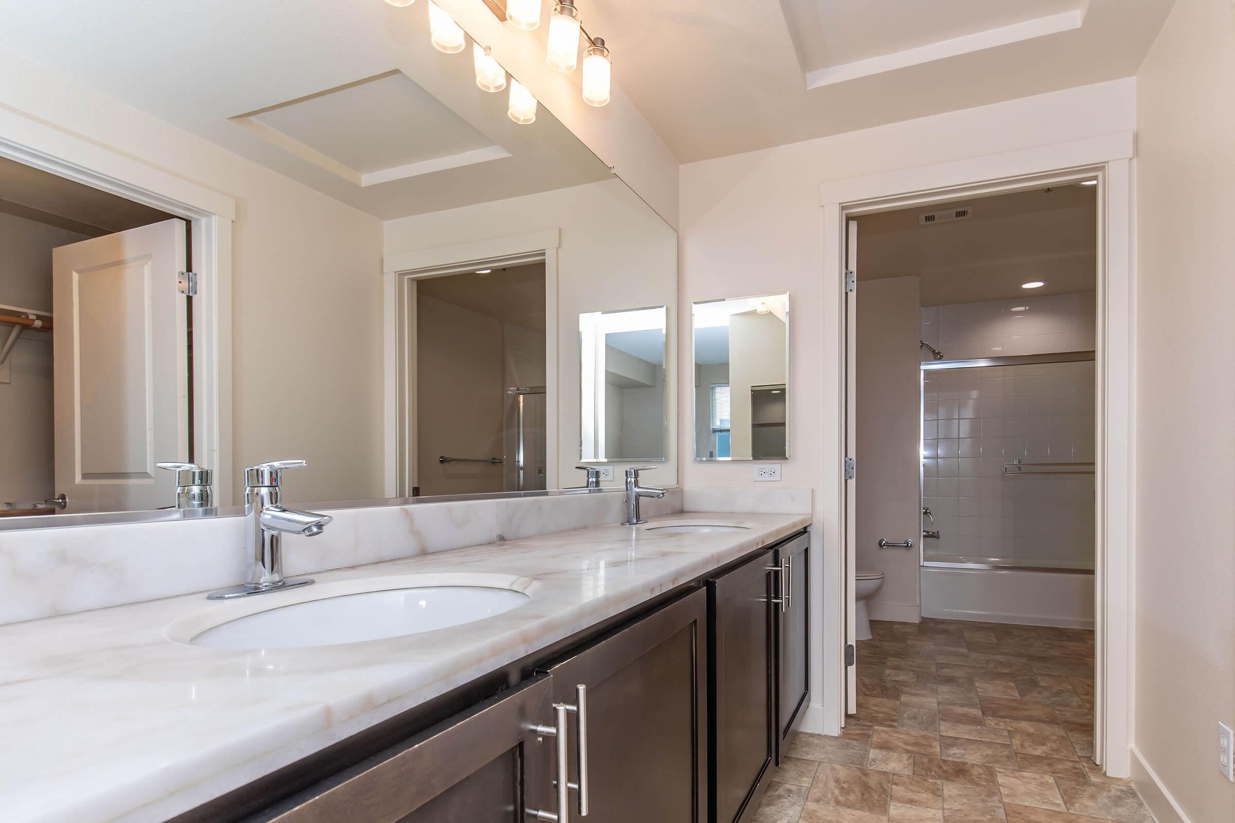 A modern bathroom featuring a double sink vanity with marble countertops, large mirrors, and a well-lit area. The space includes beige walls, a shower area in the background, and light-colored flooring, creating a clean and spacious ambiance. Two doorways lead to separate areas.