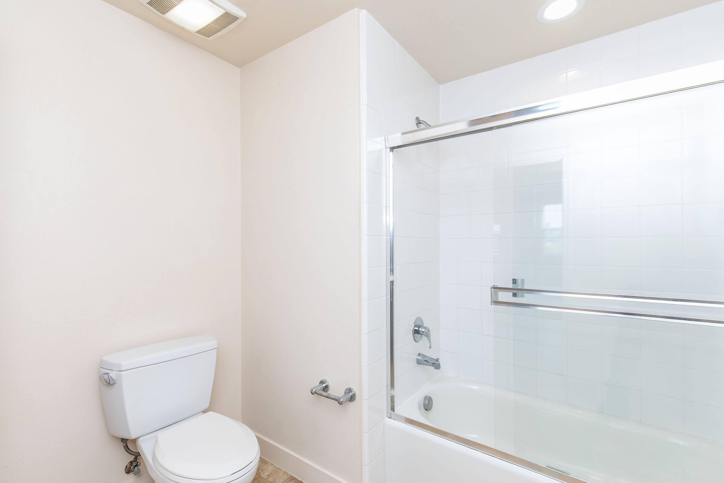 A clean, modern bathroom featuring a white toilet and a shower-tub combo with a glass sliding door. The walls are painted a light color, and there are neutral tile floors. Soft lighting from overhead fixtures illuminates the space, creating a bright and fresh atmosphere.