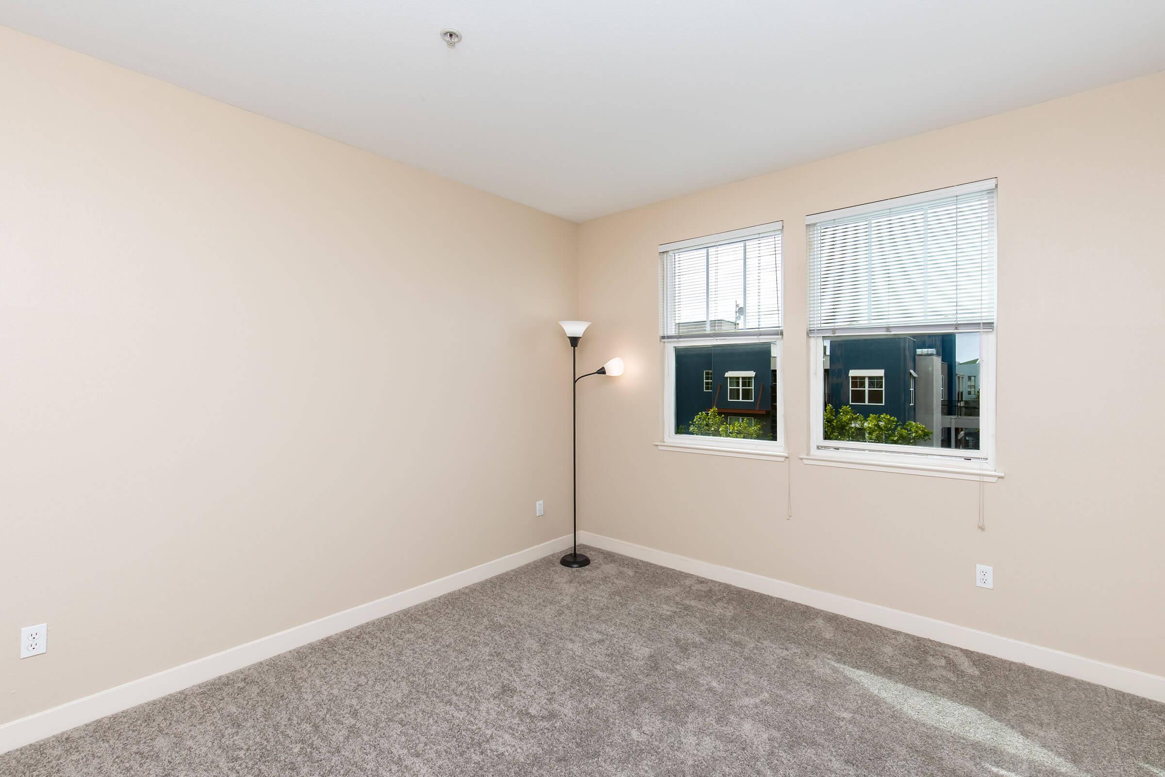 Empty room with light beige walls and gray carpet. Two windows allow natural light to enter, with a view of buildings outside. A standing lamp is positioned near one window. The space is unfurnished, creating an open and airy feel.