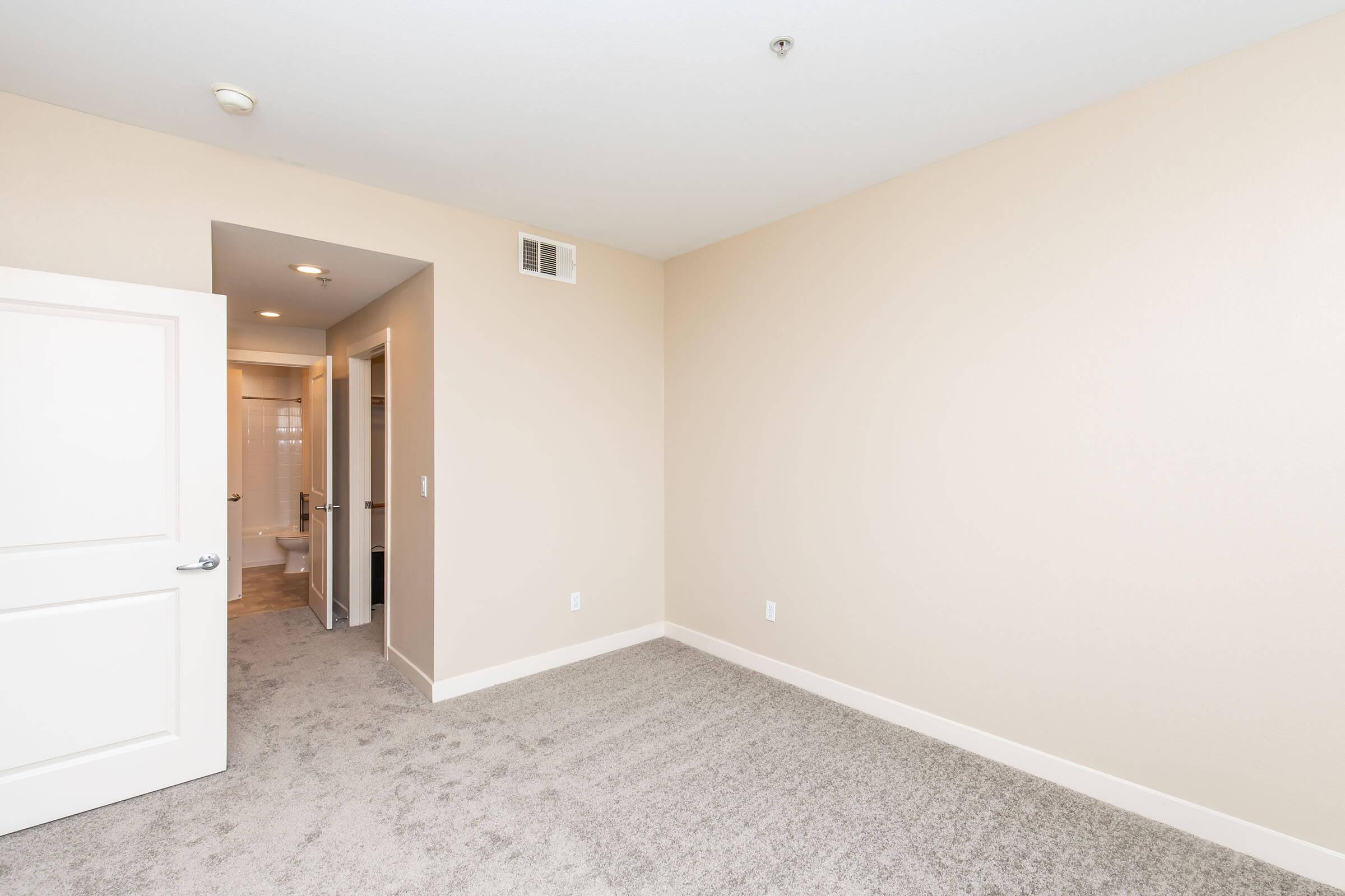 A light-colored room with carpeted flooring and neutral walls. There's a doorway leading to a bathroom, visible through an open door. The space is empty, providing a clean, minimalist aesthetic, suitable for various uses. Soft lighting enhances the airy feel of the room.